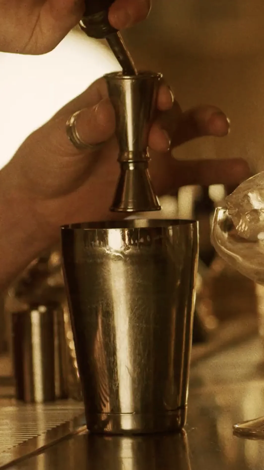 Hand pouring liquid from a jigger into a metal cocktail shaker on a bar counter.