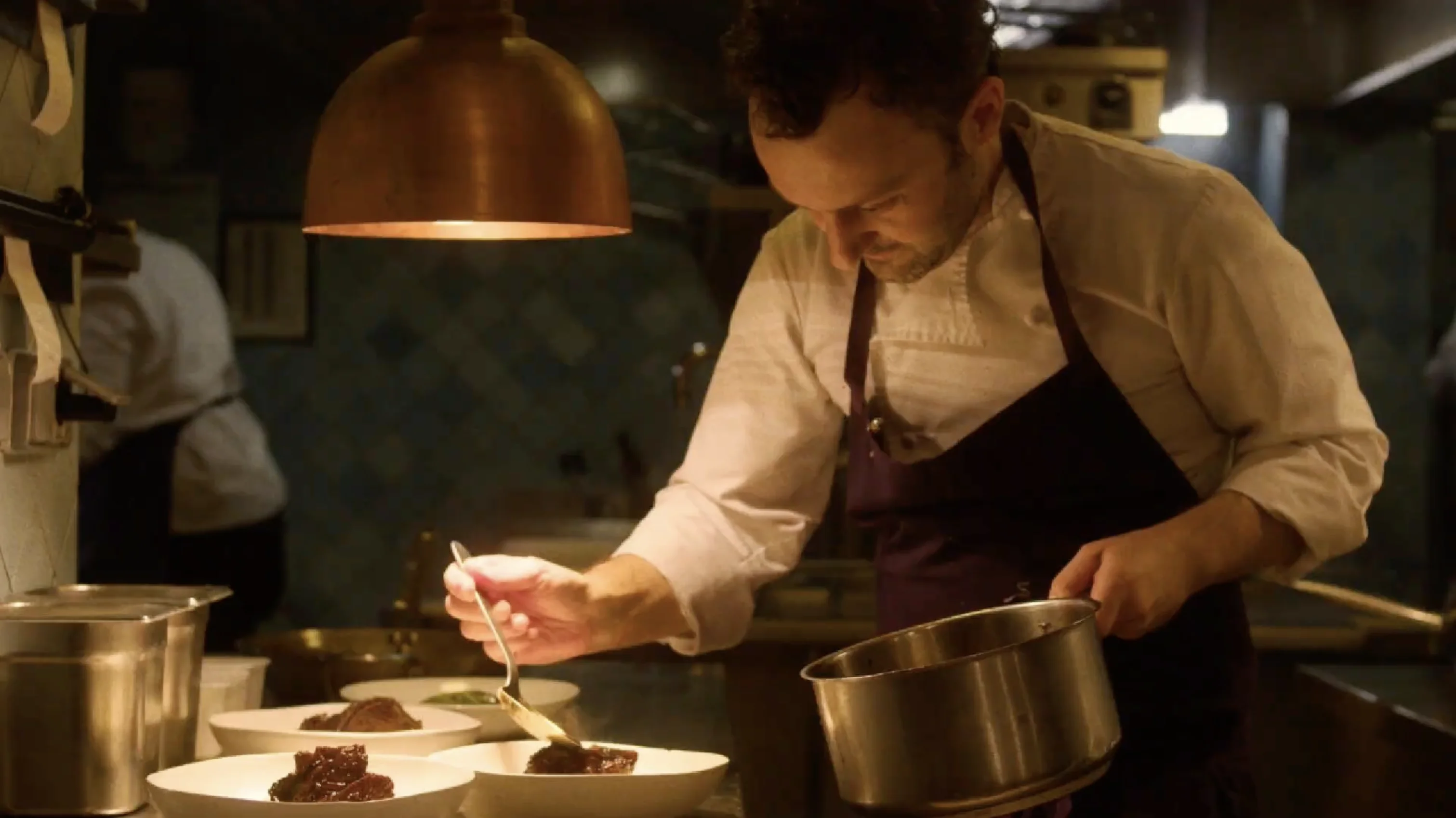 Chef in white shirt and dark apron carefully plating food under warm kitchen lighting.