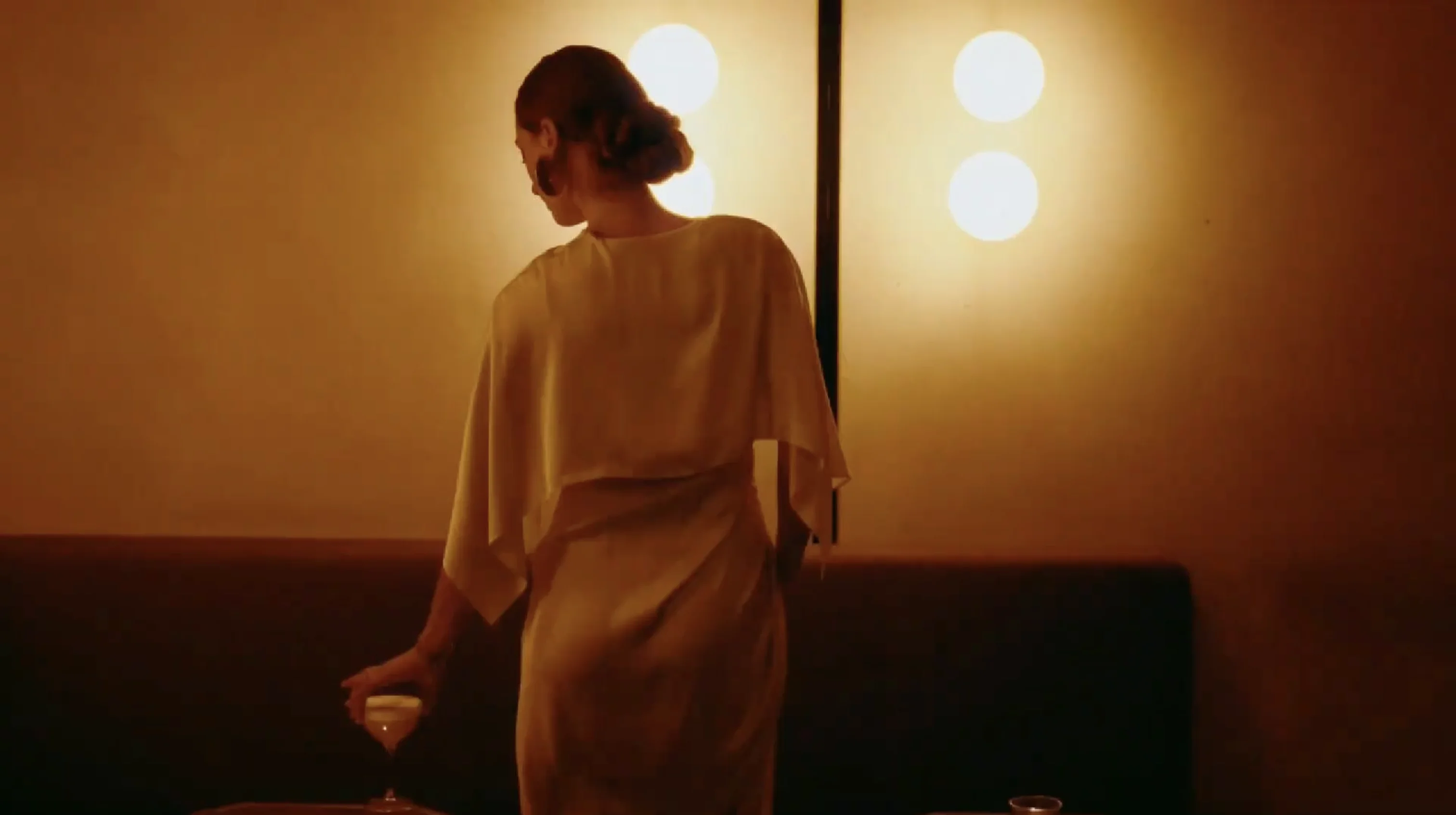 Woman in a satin dress holding a cocktail glass in a dimly lit room with round wall lights.