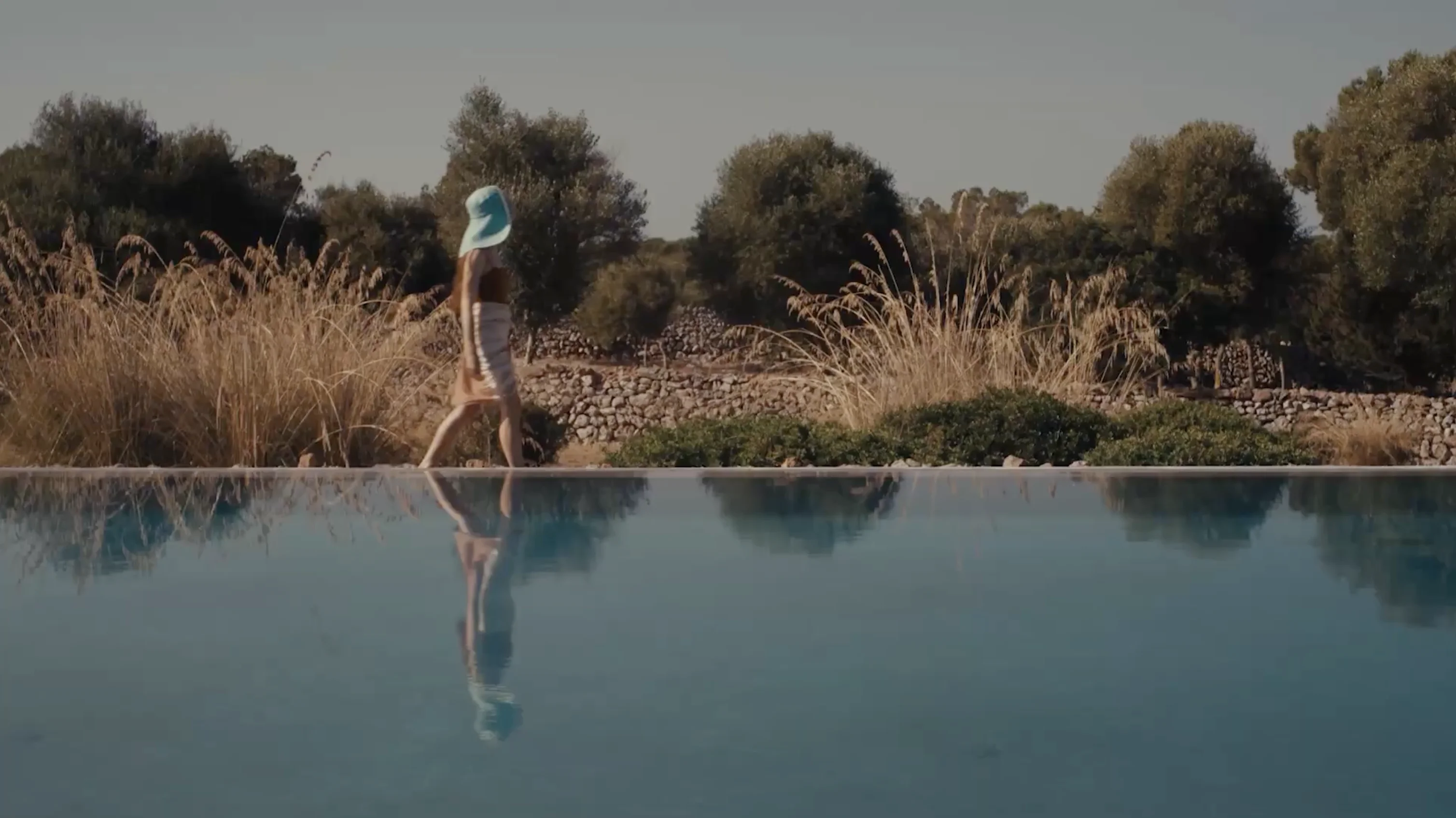 A woman wearing a blue hat walking beside a calm swimming pool with dry grass and trees in the background.