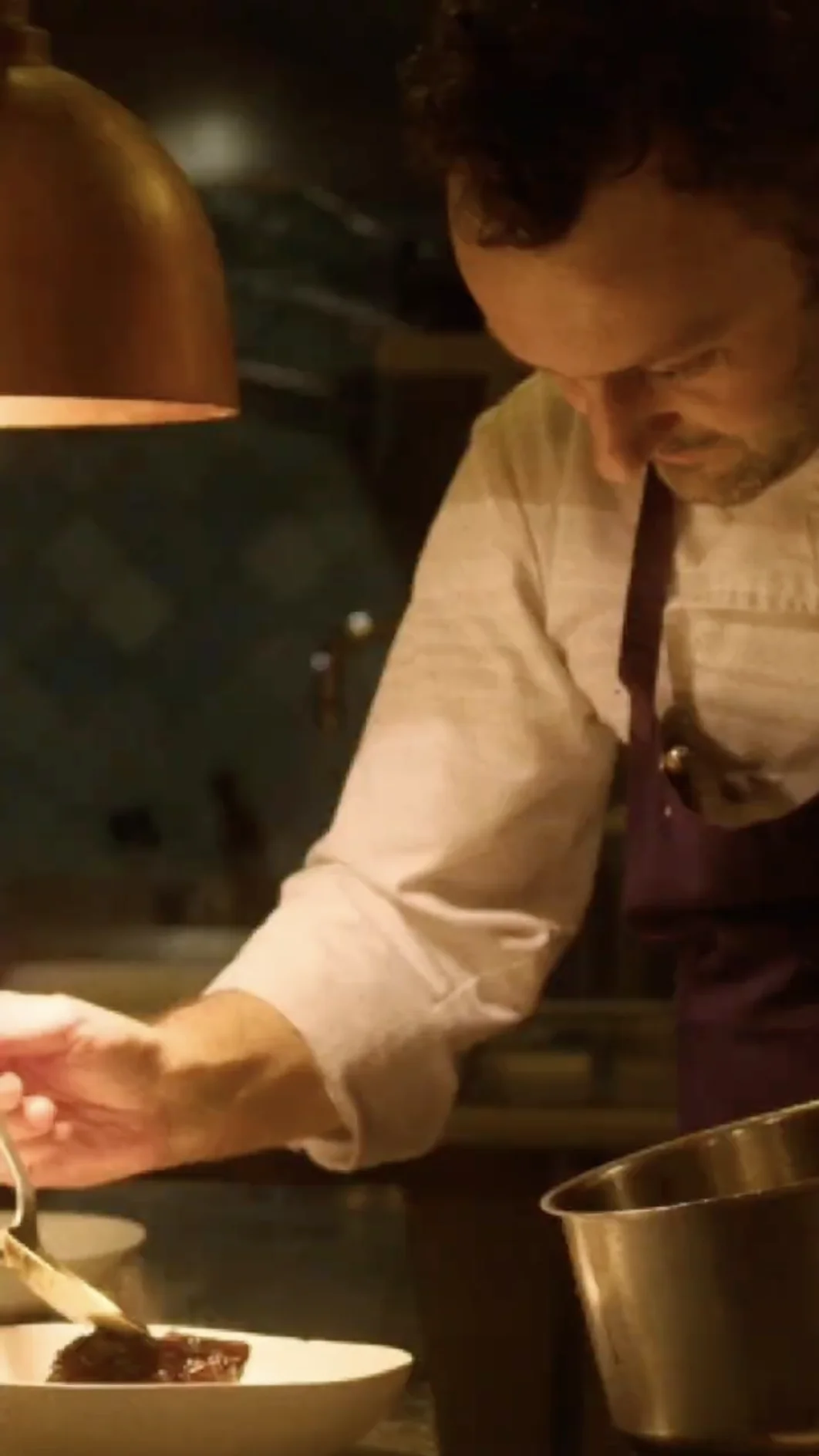 Chef wearing a white shirt and dark apron carefully plating food in a dimly lit kitchen.