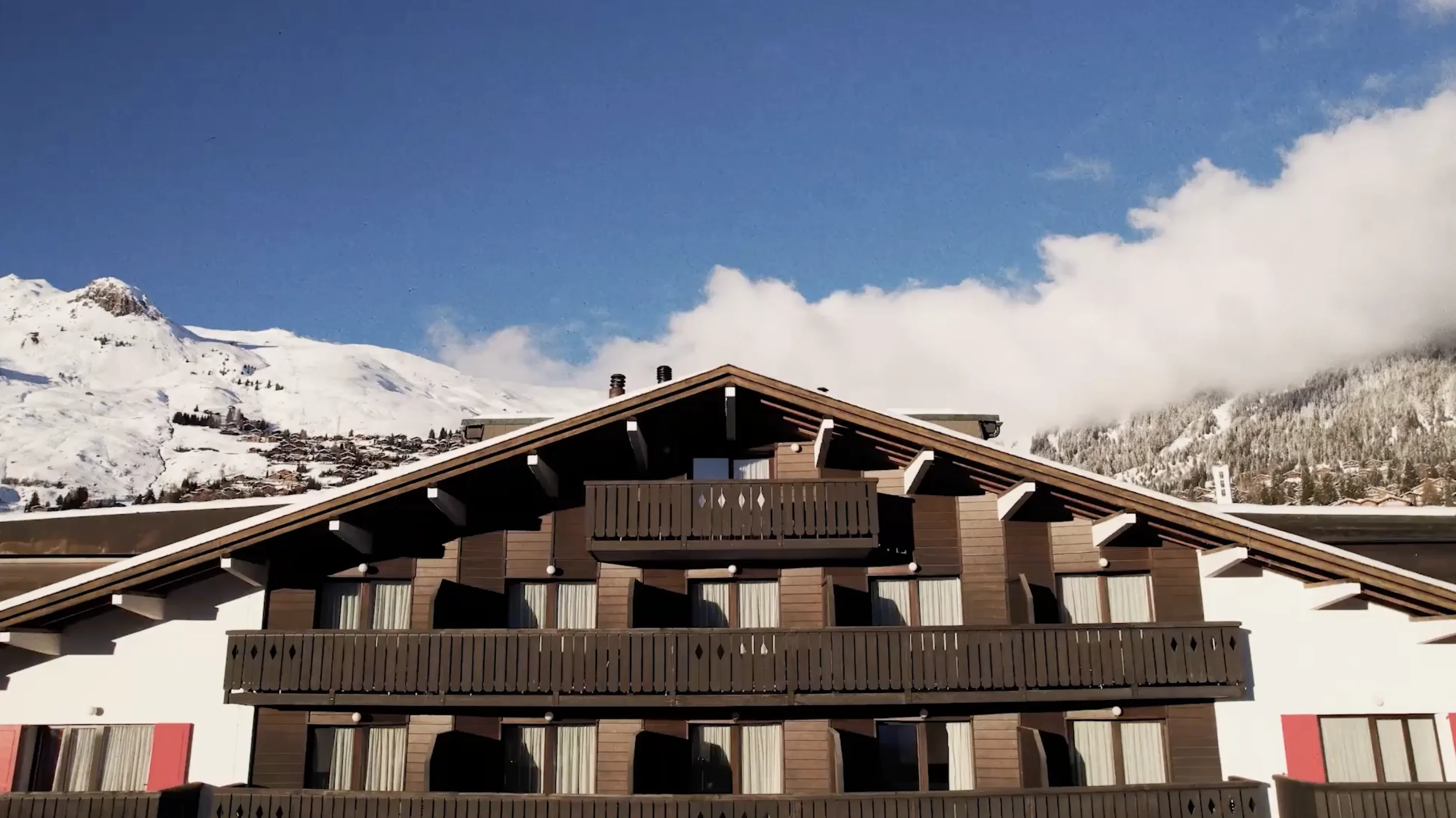 Mountain chalet with dark wooden balconies and snow-covered peaks in the background under a blue sky.