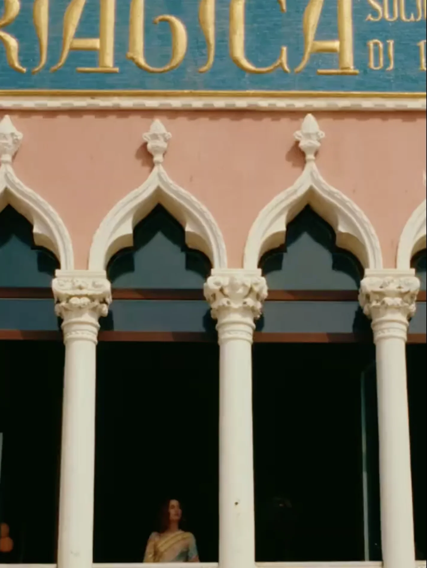 Woman in a traditional saree standing behind white ornate arches of a Venetian-style building with a peach facade.