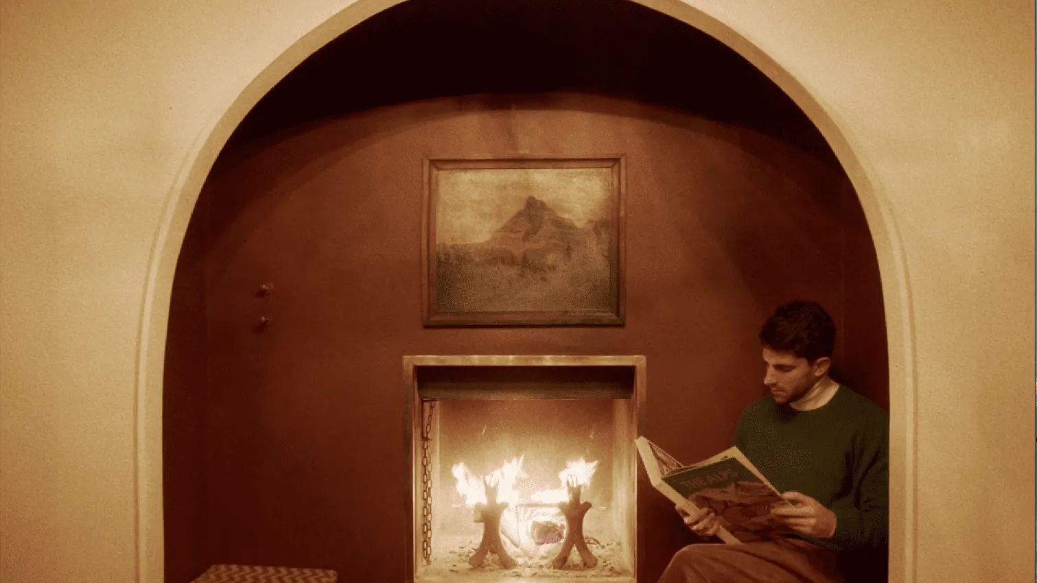 Man sitting beside a fireplace under an arched alcove, reading a book, with a painting of a mountain above the fireplace.