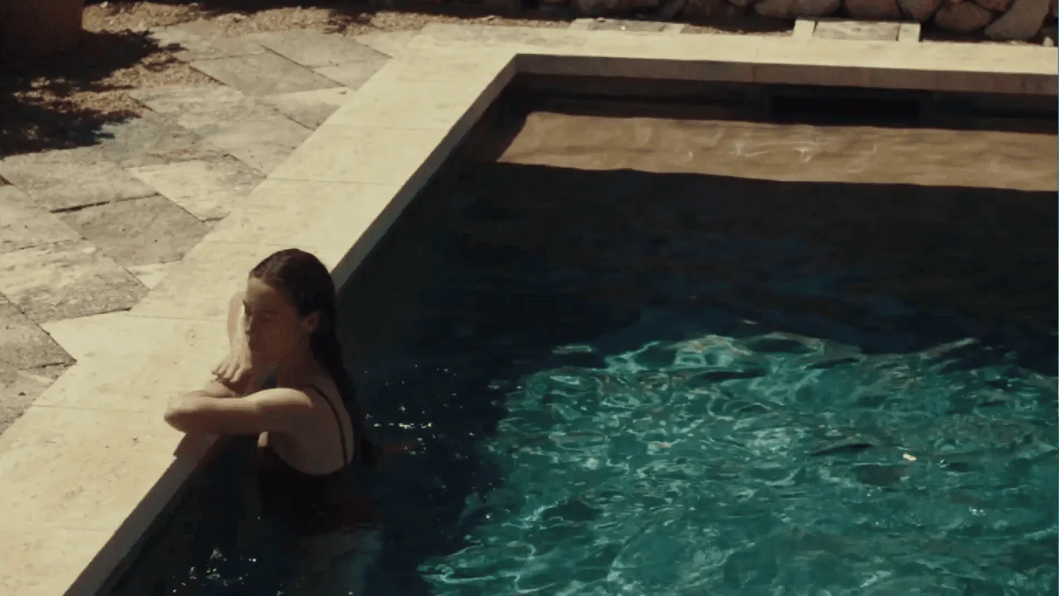 Person with wet hair leaning on the edge of a swimming pool in bright sunlight.
