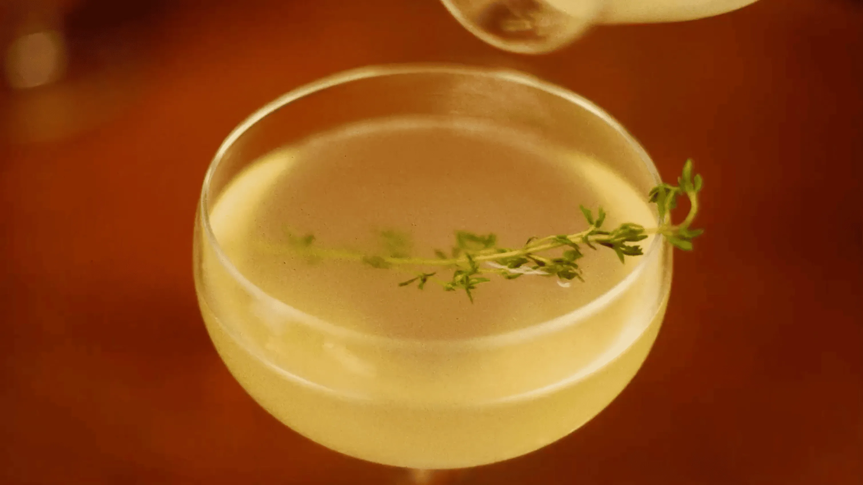 Cocktail in a coupe glass garnished with a sprig of fresh thyme on a brown surface.