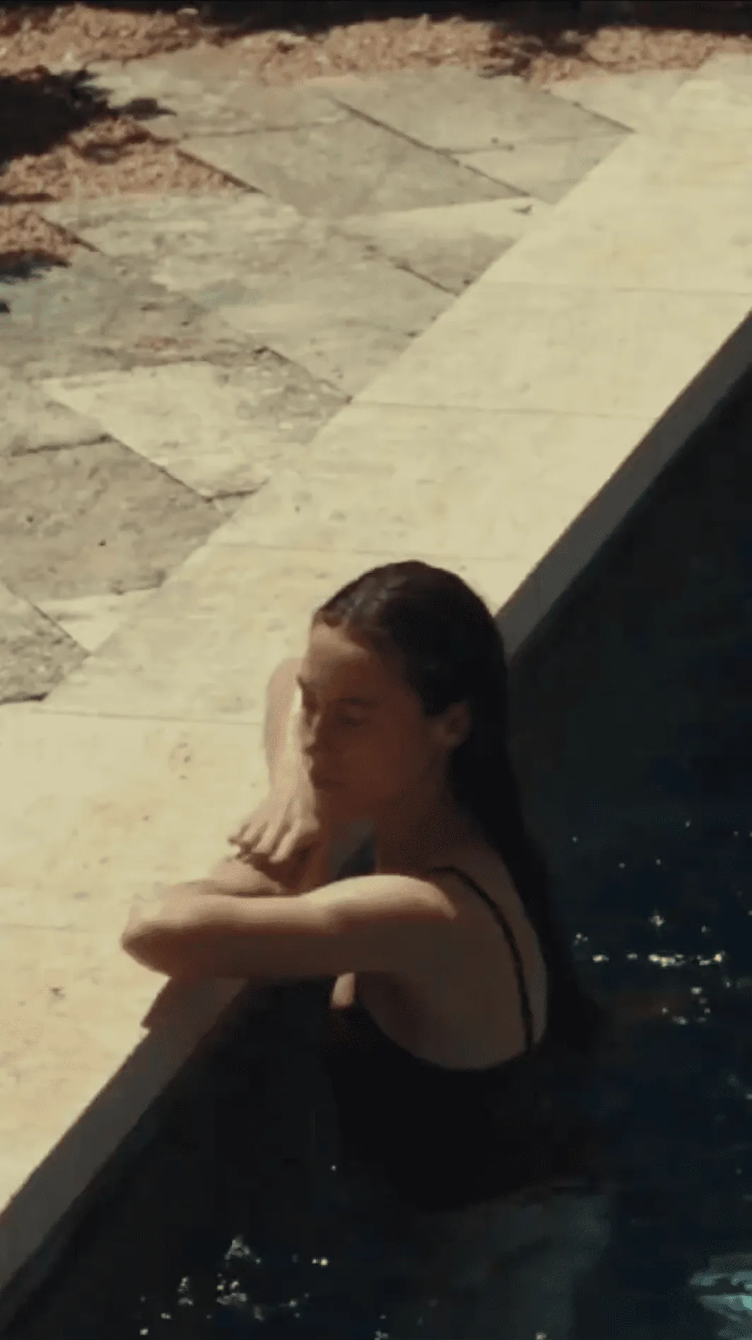 Young woman in a black swimsuit resting her arms on the edge of a pool outdoors.