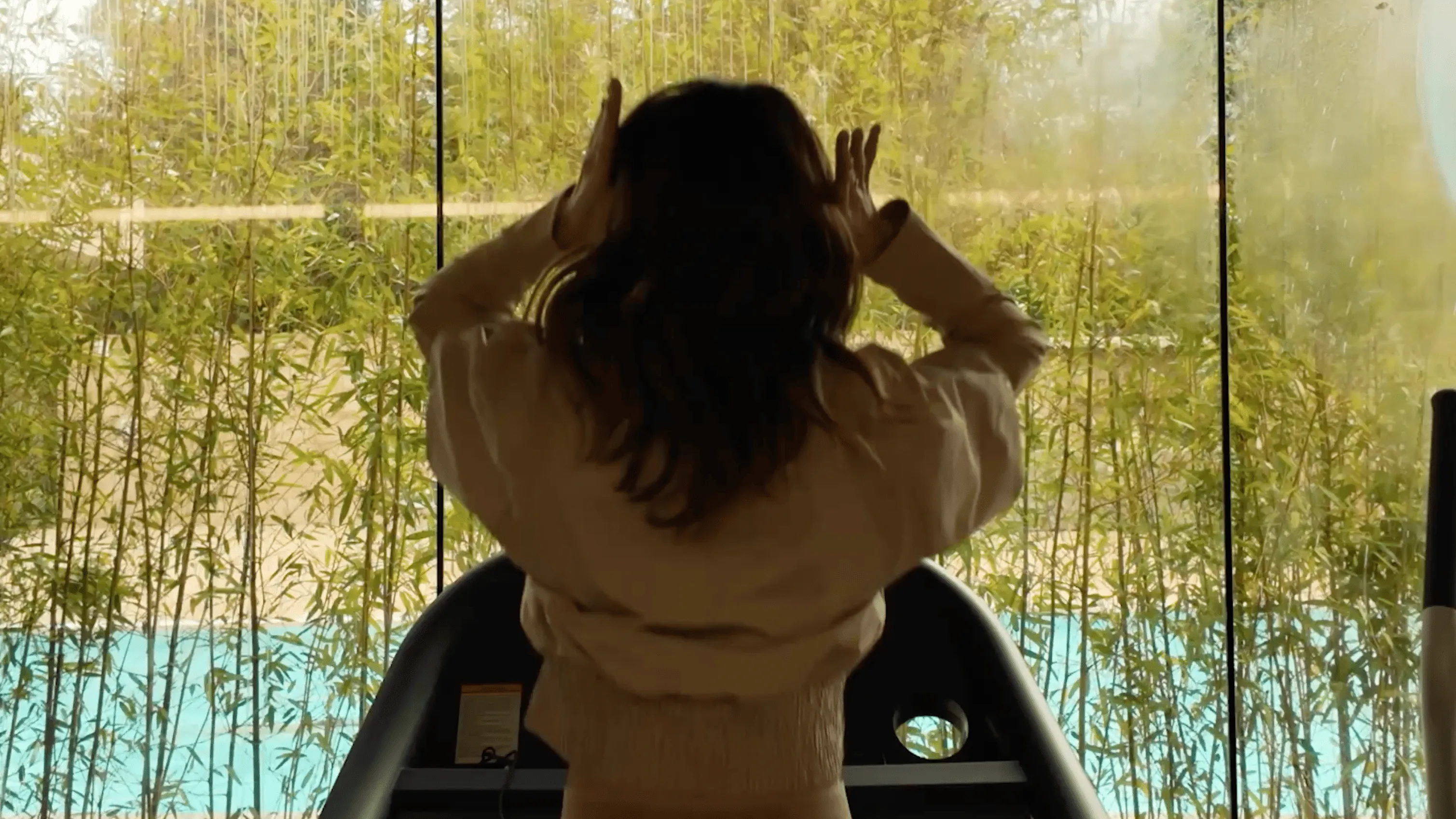 Woman with dark hair standing on a treadmill, facing a large window with green bamboo plants outside.