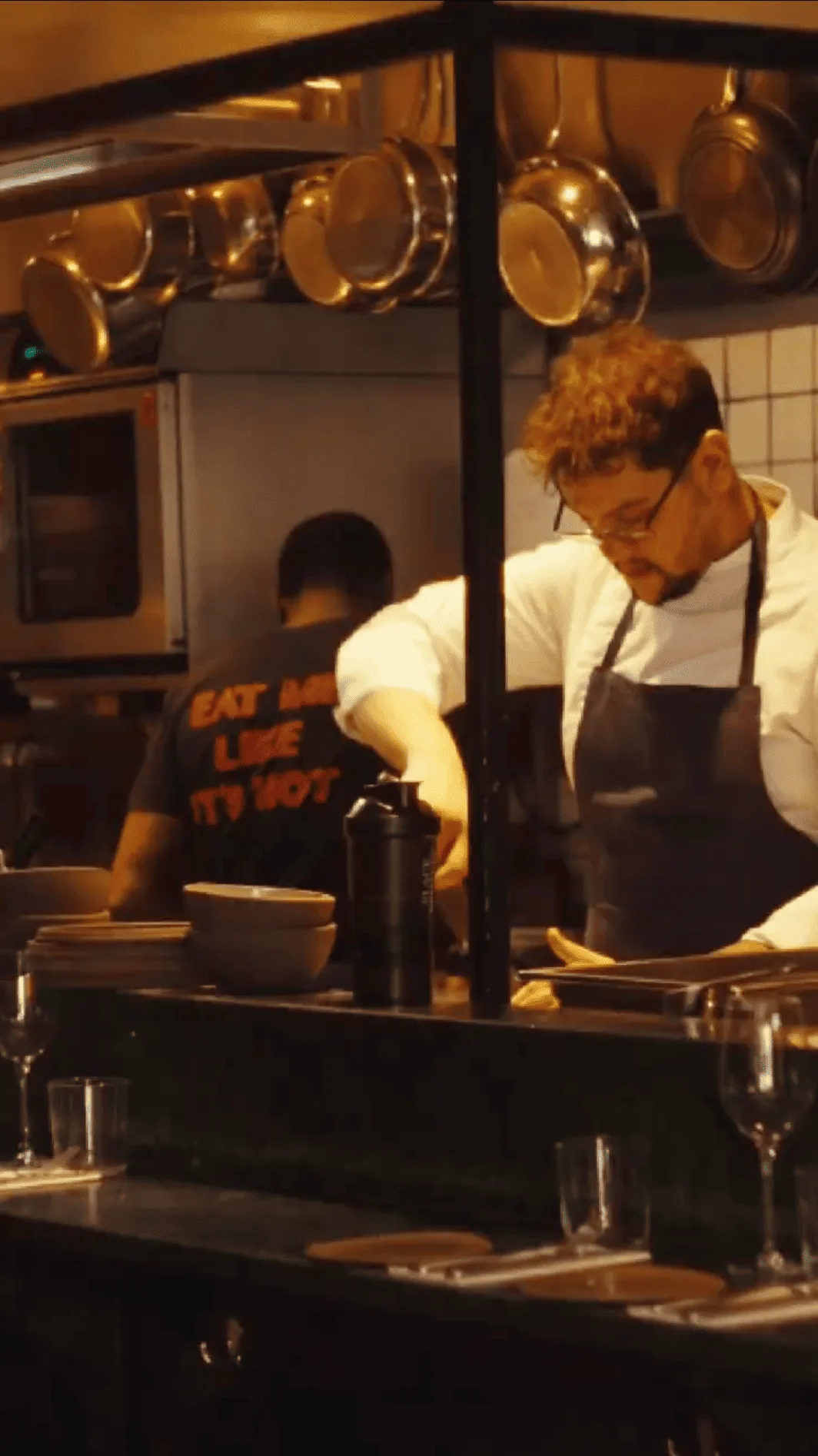 Chef wearing glasses and apron preparing food behind a counter in a kitchen with pots hanging above.