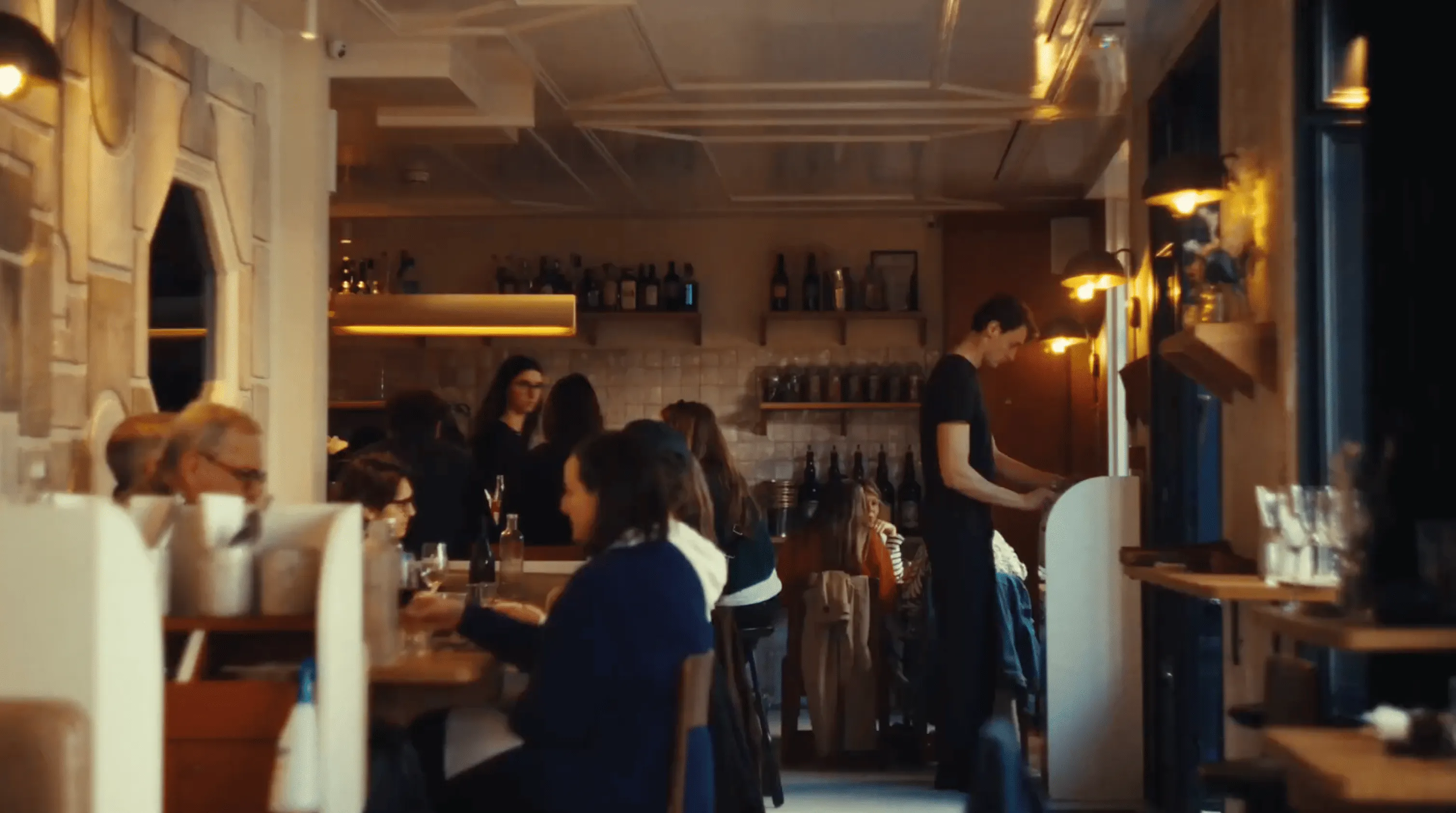Interior of a cozy restaurant with people dining and a waiter standing by a counter.