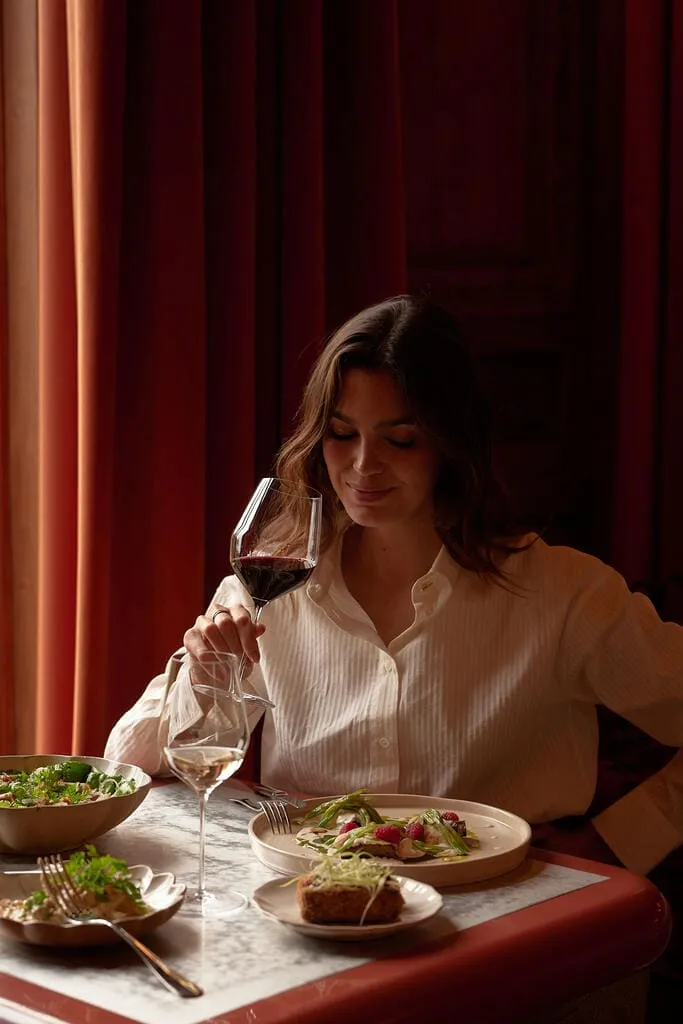 Woman in white blouse enjoying a glass of red wine while sitting at a table with plates of gourmet food and a glass of white wine.