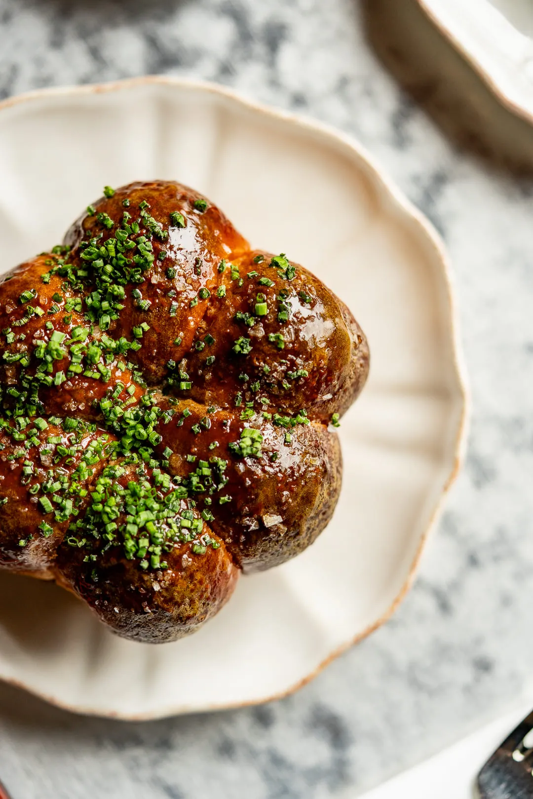 Glazed brown bread roll garnished with chopped chives on a white scalloped plate.