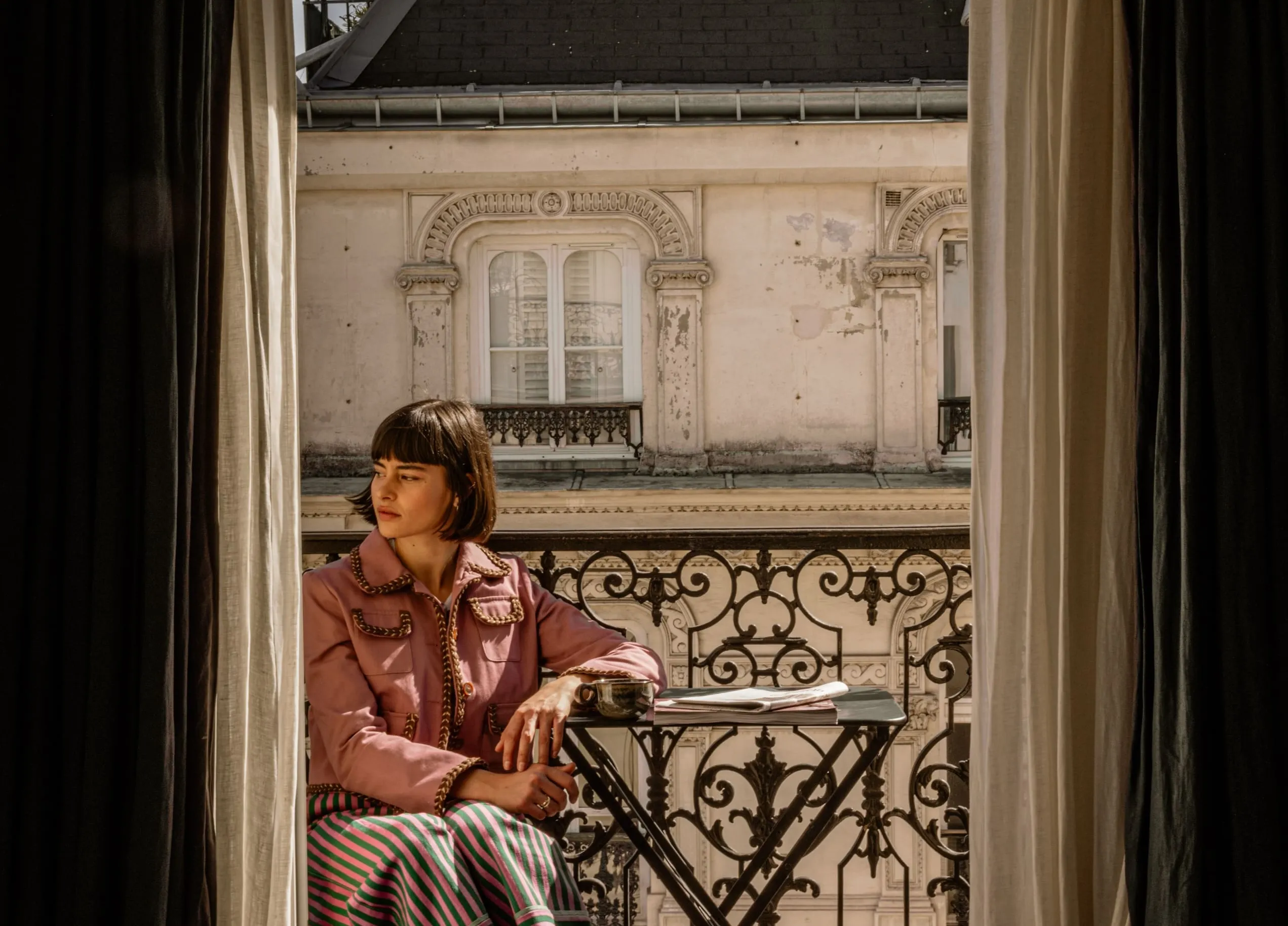 Femme dans une veste rose assise sur un balcon avec une balustrade en fer ornée, regardant sur le côté avec une tasse et un journal sur une petite table.
