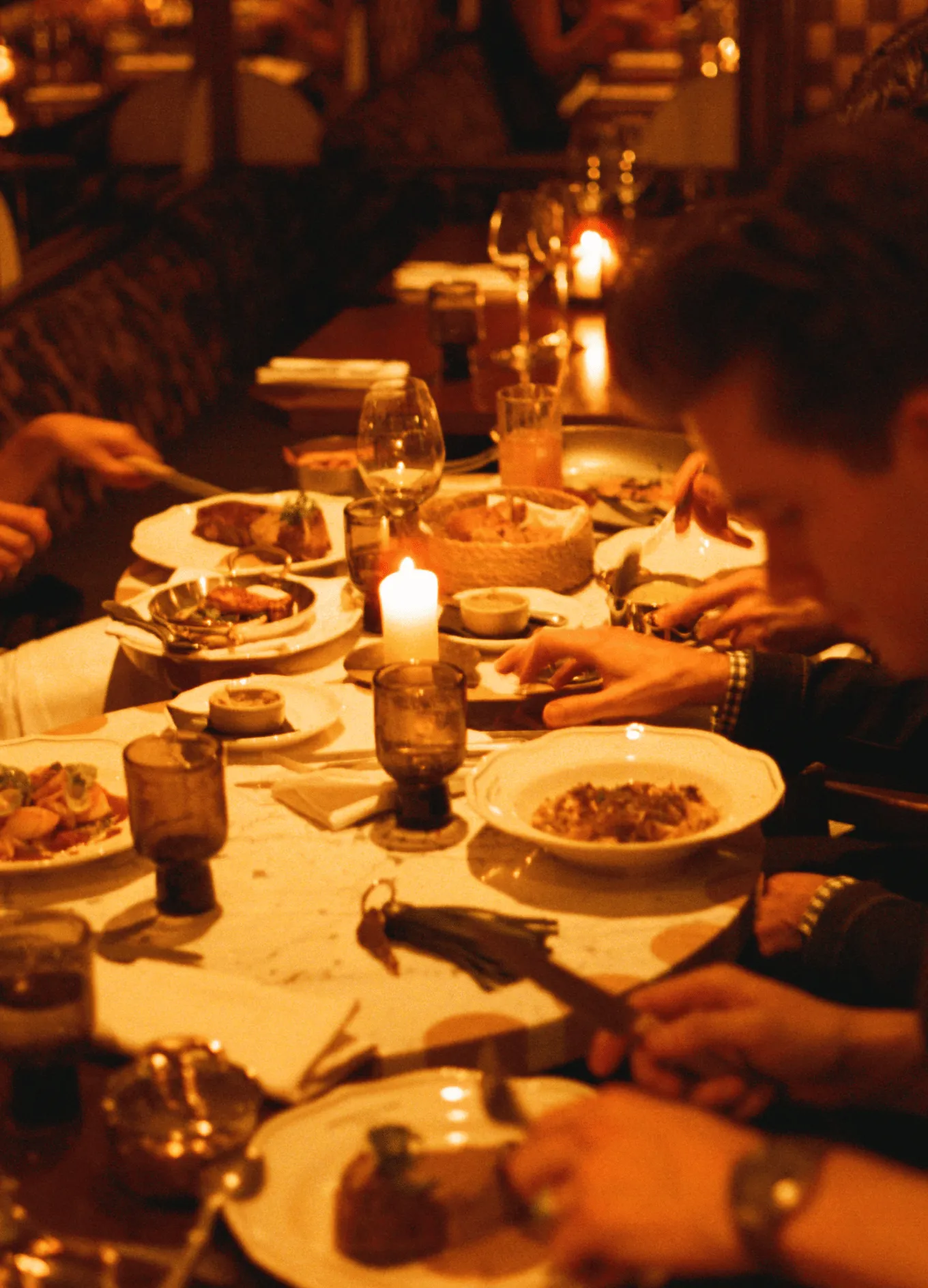 People dining at a warmly lit table with candles and various plated dishes in a cozy restaurant setting.