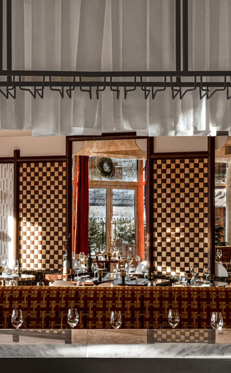 Elegant restaurant interior with patterned walls, large windows showing outdoor greenery, and a marble countertop with empty wine glasses.
