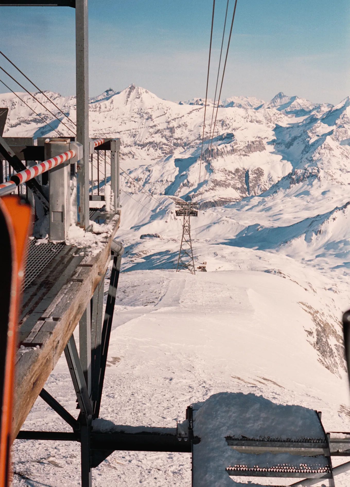 Snow-covered mountain landscape with a ski lift tower and cables extending into the distance under a clear blue sky.