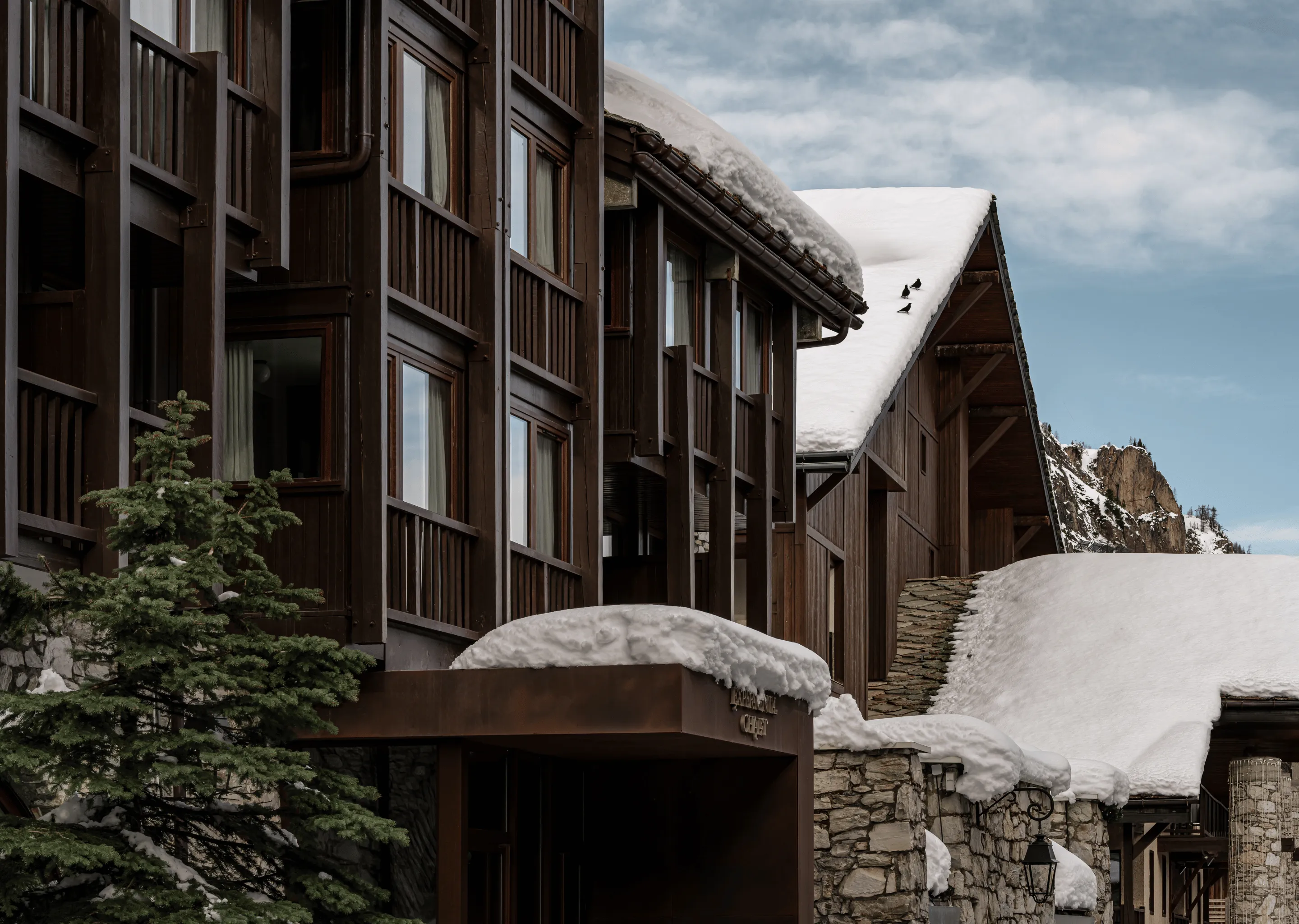 Wooden and stone chalet covered in snow with a snow-capped roof and pine tree in front under a cloudy blue sky.