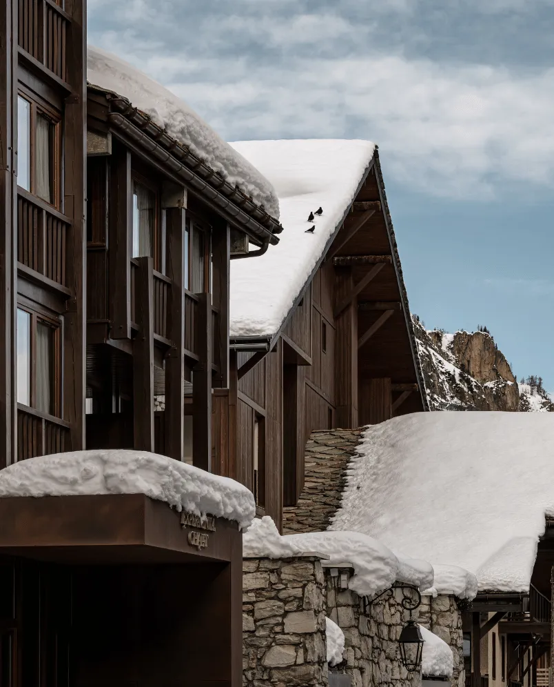 Snow-covered chalet with wooden exterior and stone walls under a cloudy sky and mountain backdrop.