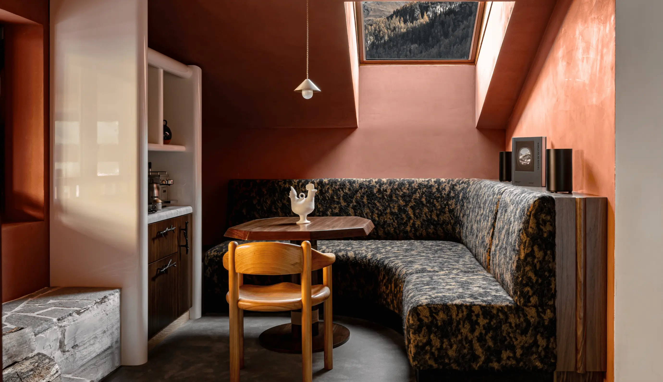 Cozy corner dining area with patterned curved bench, wooden chair, wooden table, skylight window, and terracotta walls.