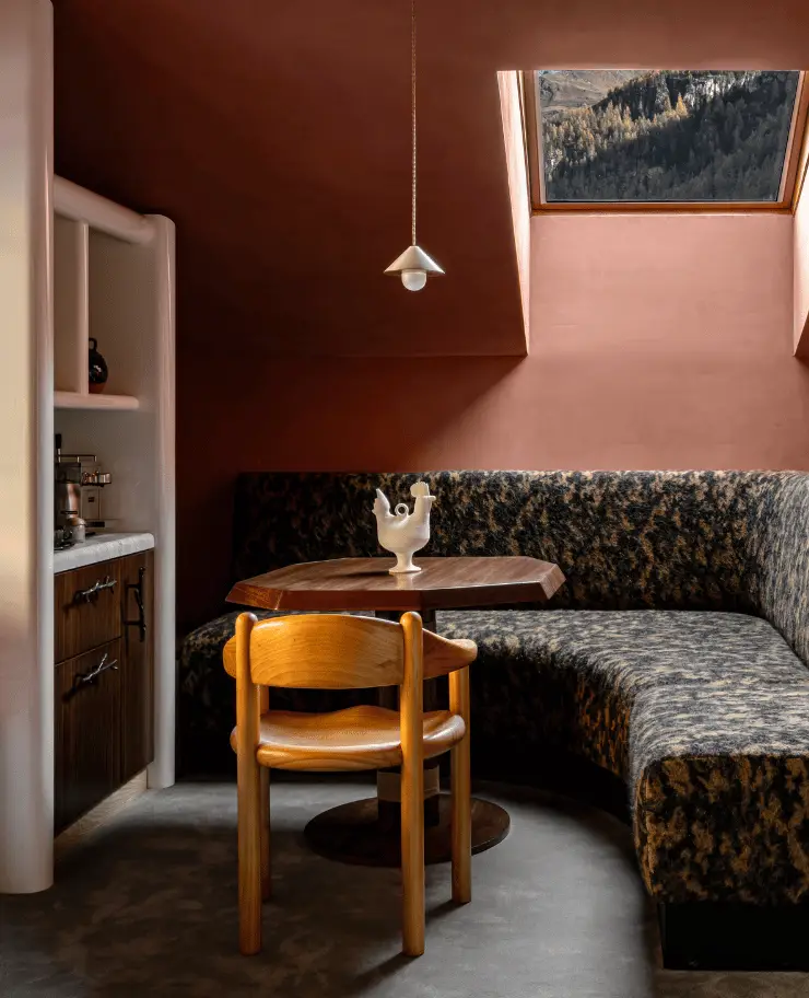 Cozy corner seating with patterned bench, wooden chair, octagonal wooden table, hanging light, and skylight window showing mountain trees.