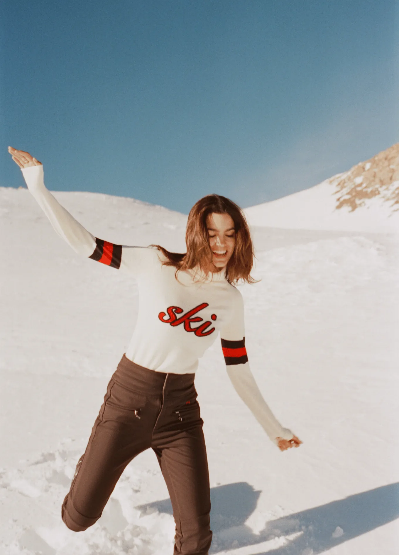Smiling woman wearing a white sweater with the word 'ski' and brown pants, posing joyfully on snowy mountains under a clear blue sky.