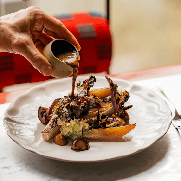Hand pouring sauce onto a gourmet dish with grilled vegetables and mushrooms on a white plate.