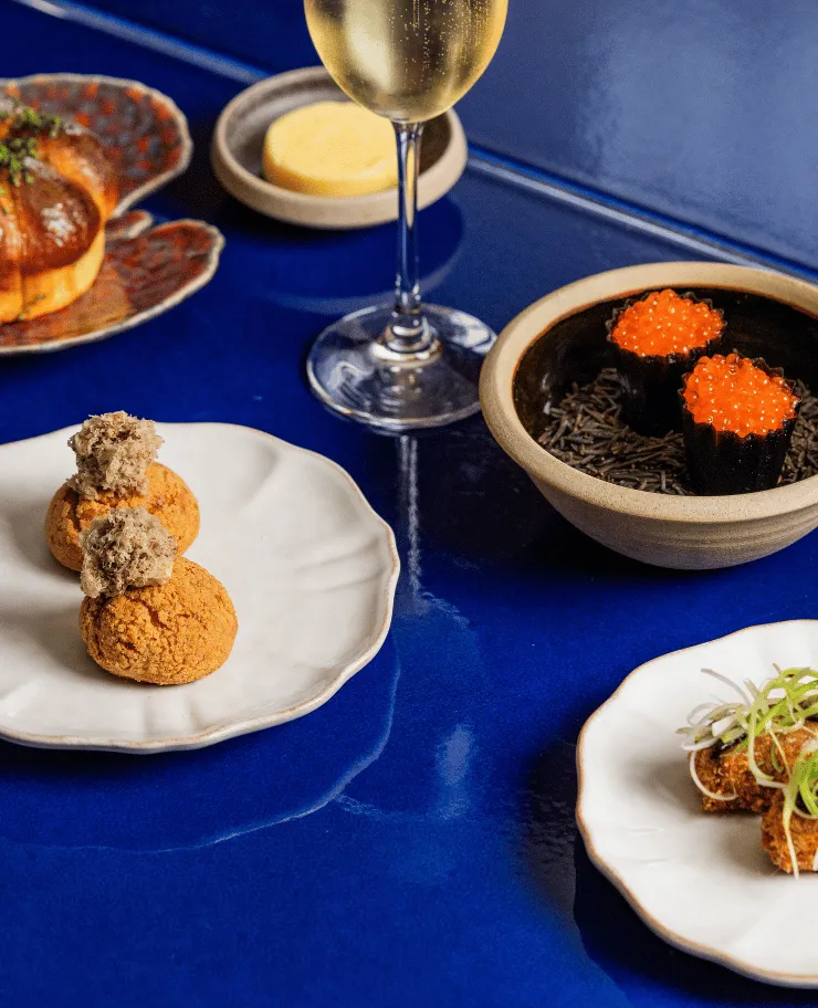 Elegant table setting with gourmet appetizers including two small puffs with a topping, sushi with bright orange roe in a bowl, and a glass of sparkling wine on a deep blue glossy surface.