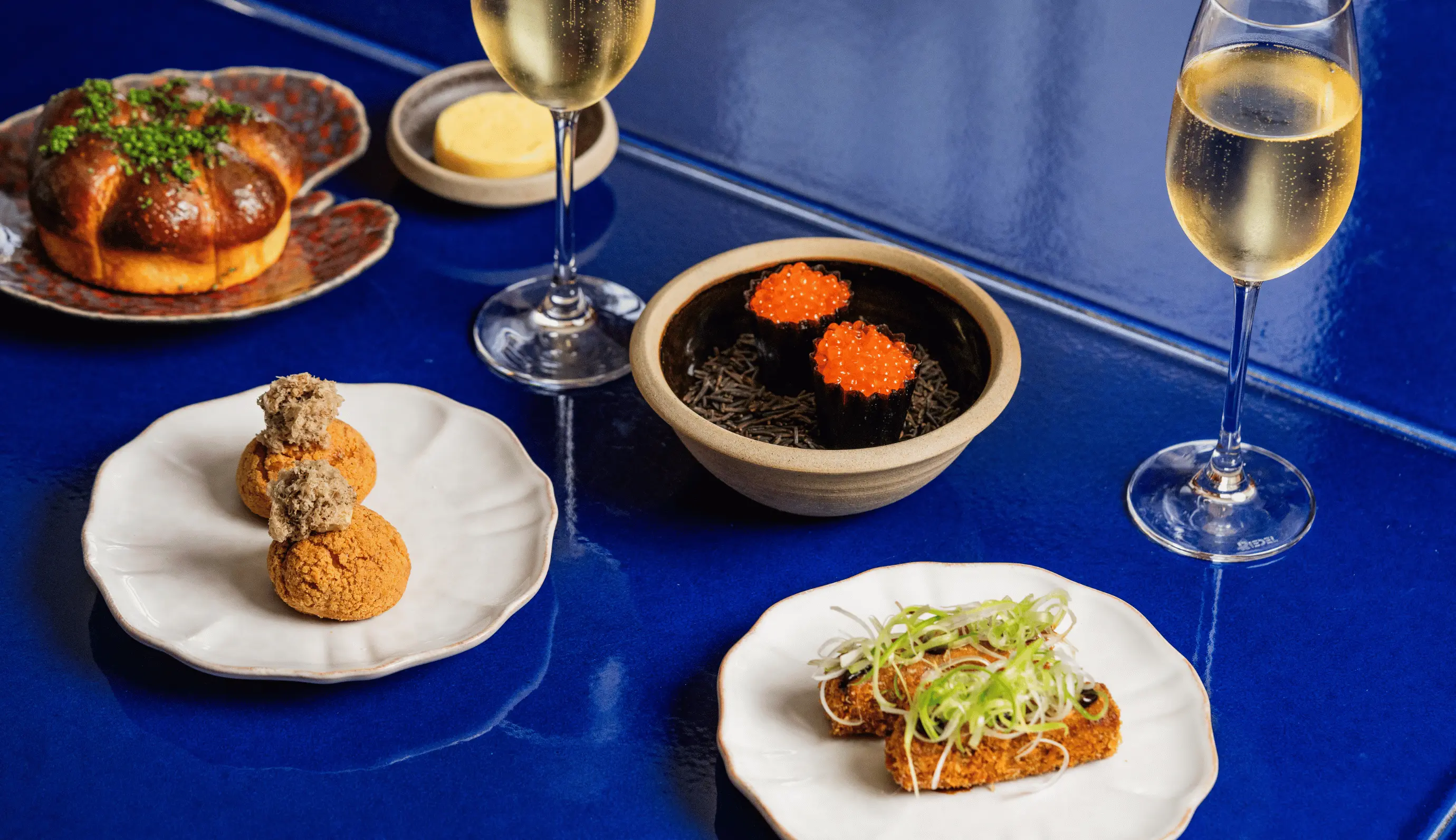 Gourmet dish assortment on a blue table, including two glasses of sparkling wine, a mushroom-shaped bread with herbs, two round pastry puffs topped with a mousse, two sushi cups with orange fish roe, and two fried pieces garnished with sliced green onions.