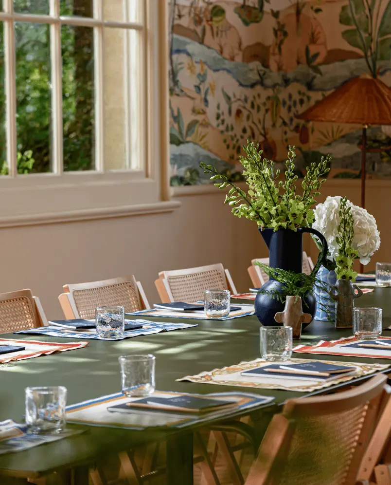 Long green table set for a meeting or meal with notebooks, glasses, placemats, and flower arrangements in a well-lit room with a large window and wallpaper.