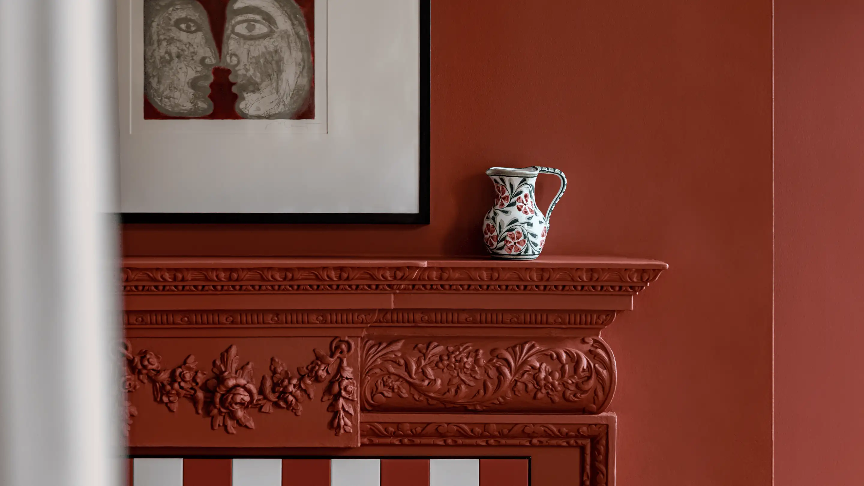 Decorative red fireplace mantel with floral carvings, a white pitcher with red flowers on top, and artwork of two faces on the wall.