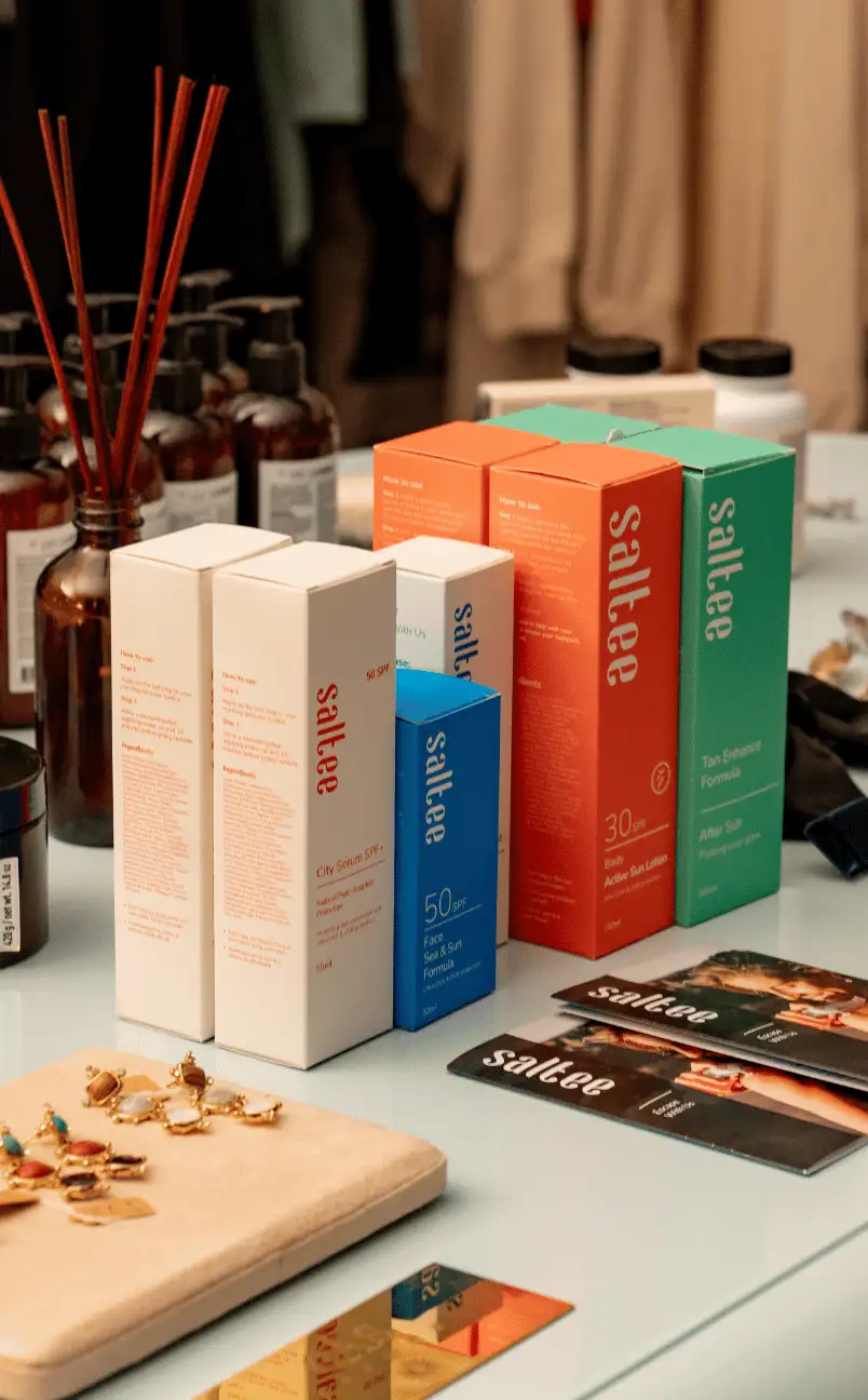 A variety of Saltee skincare product boxes including face and sun formulas displayed on a table with jewelry and brown bottles in the background.