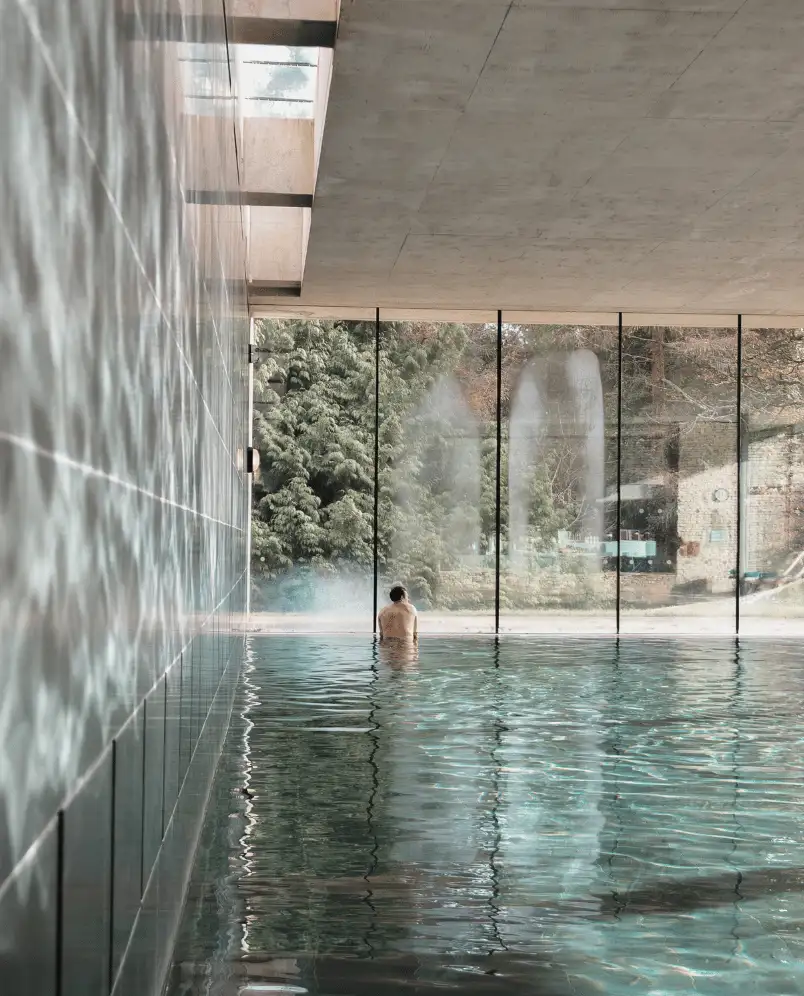 Person sitting in an indoor pool facing large floor-to-ceiling windows overlooking greenery outside.