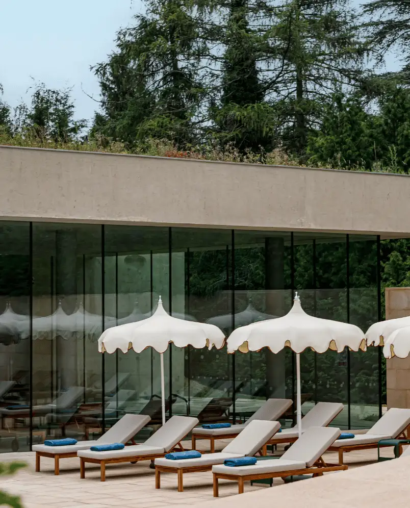 Modern outdoor lounge area with wooden sun loungers topped with gray cushions and blue towels, shaded by white scalloped umbrellas in front of a glass building and surrounded by greenery.