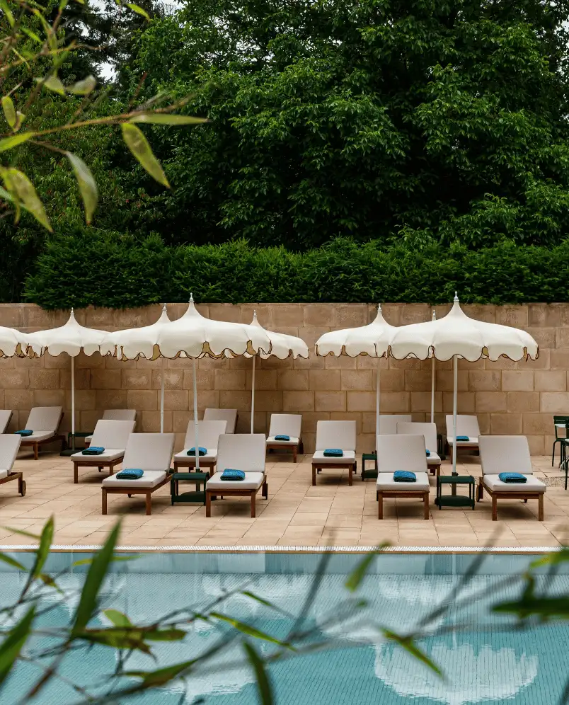 Rows of white lounge chairs with folded blue towels under cream umbrellas by a tiled poolside with green trees and hedge backdrop.