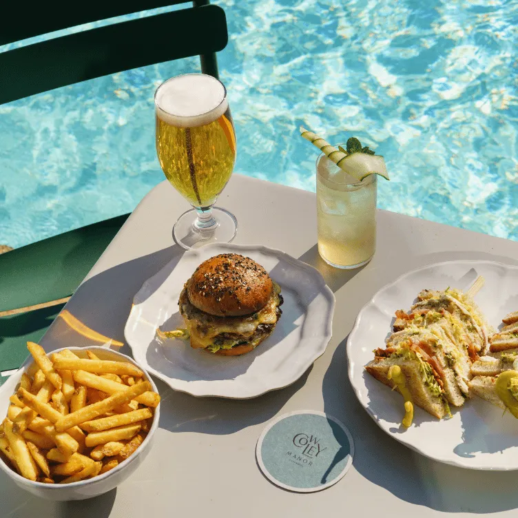 Table by pool with a cheeseburger, french fries, sandwich slices, a glass of beer, and a cocktail garnished with cucumber and mint.