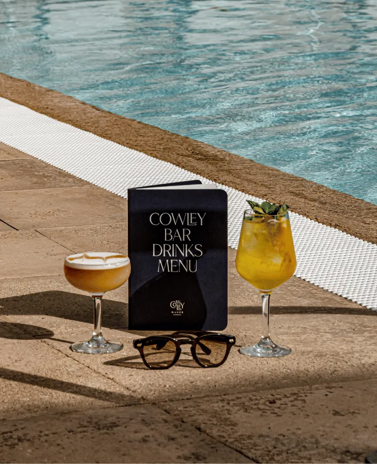 Two cocktails and a black drinks menu labeled 'Cowley Bar Drinks Menu' placed on the poolside pavement next to a pair of tortoiseshell sunglasses.