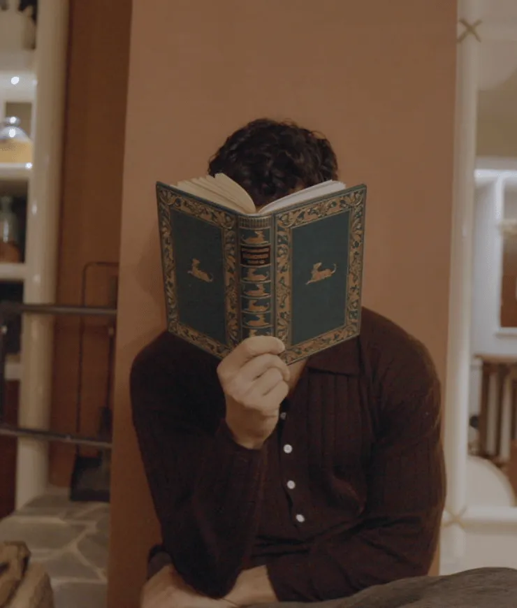 Person sitting indoors holding an ornate green book with gold designs, covering their face.