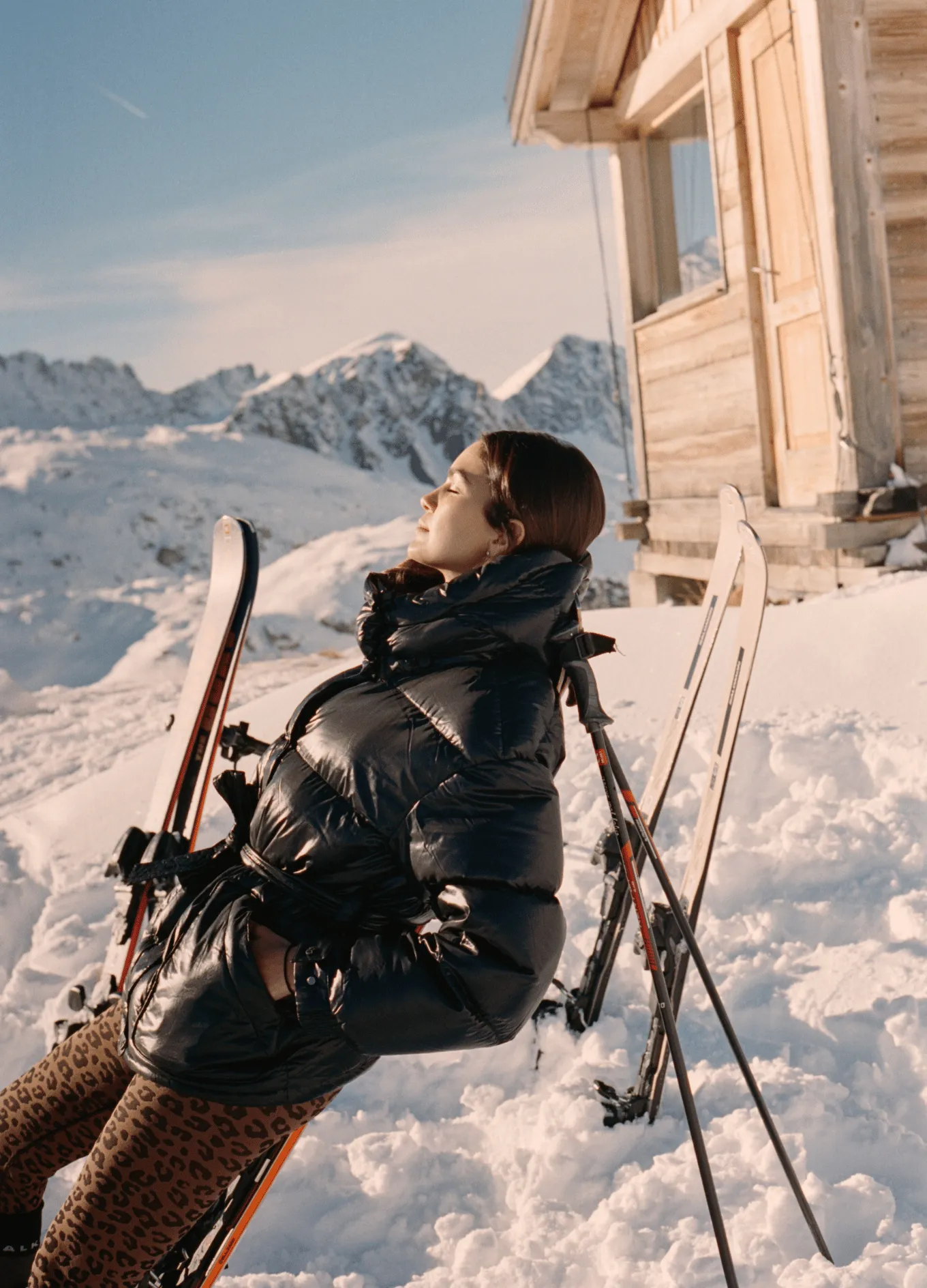 Woman in a black puffer jacket leaning back on skis in snowy mountains near a wooden cabin, eyes closed in sunlight.
