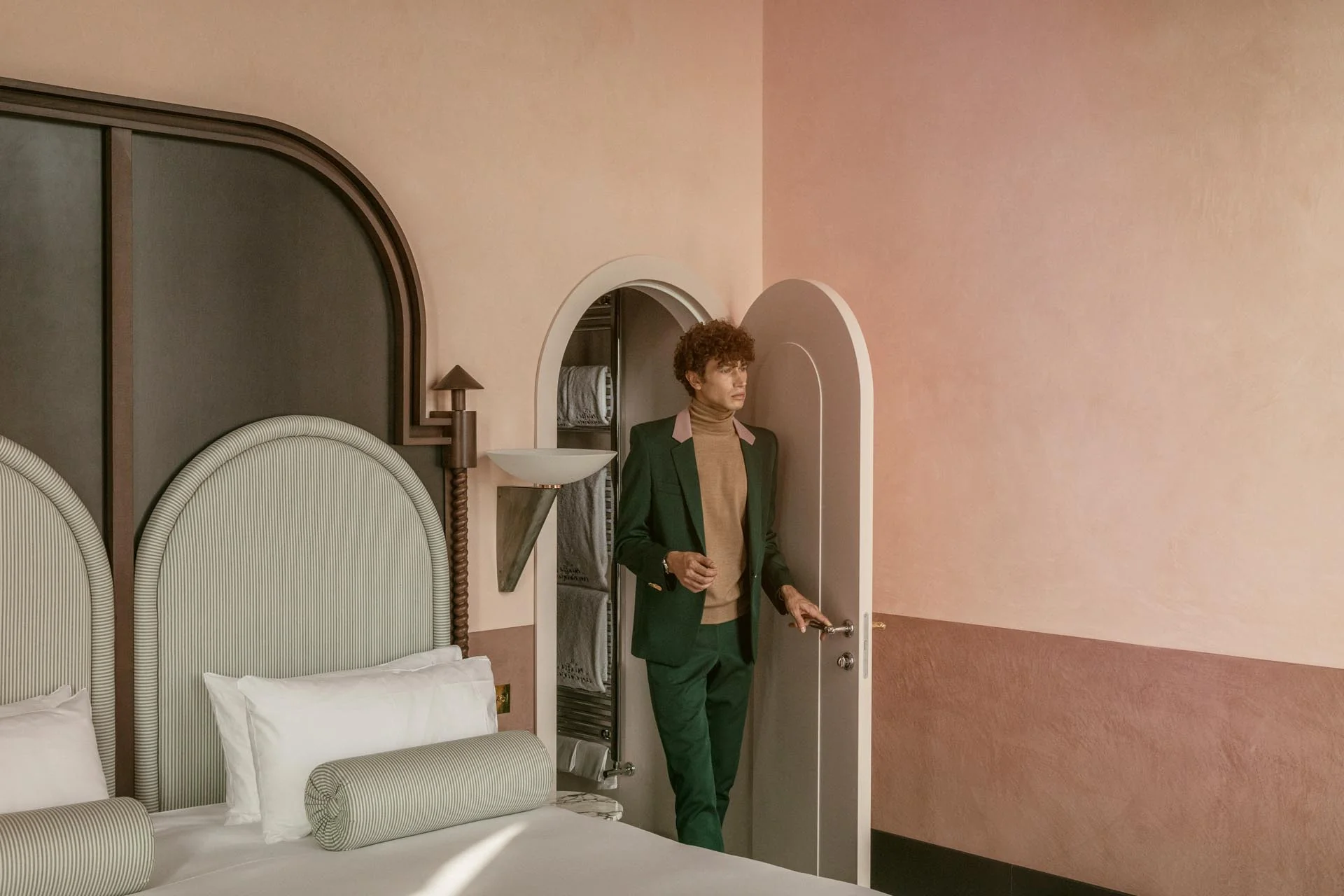 Homme en costume vert et col roulé beige ouvrant une porte cintrée dans une chambre à coucher avec une tête de lit à rayures blanches et des murs roses.