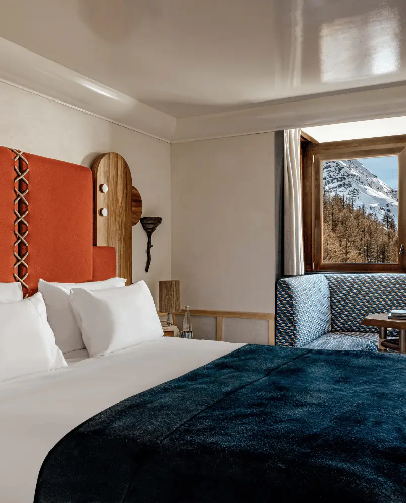 Cozy hotel room with a large bed featuring white pillows and a dark blue blanket, an orange headboard, a wooden wall lamp, and a window showing snowy mountains and trees.