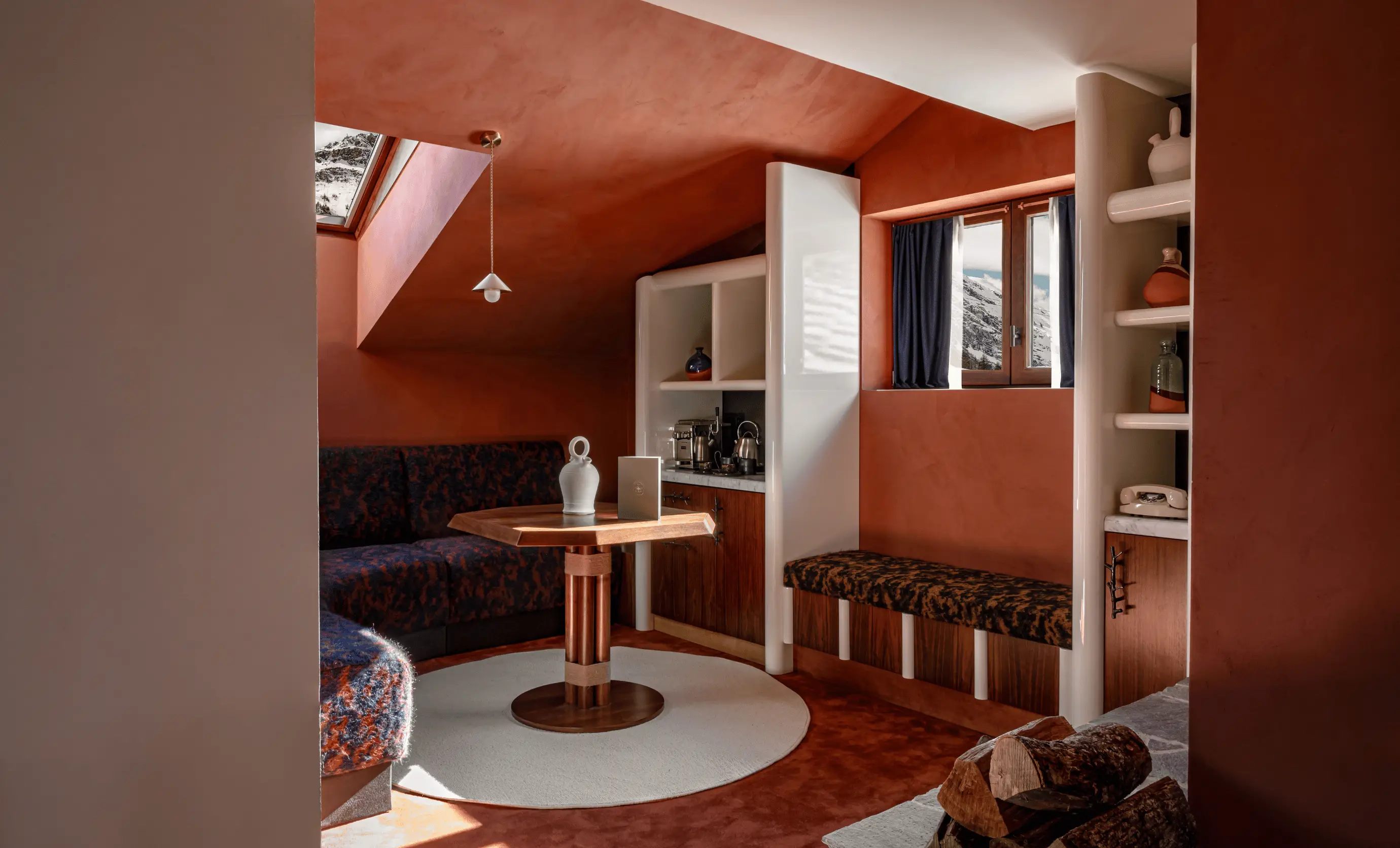 Cozy hotel room corner with burnt orange walls, built-in seating with patterned upholstery, a wooden table, and a window showing a snowy mountain view.