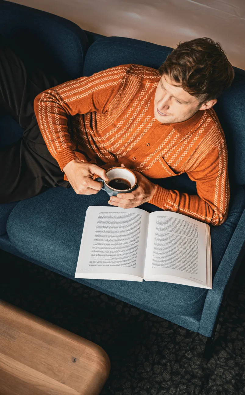 Man in an orange patterned sweater lying on a blue couch holding a cup of black coffee with an open book in front of him.