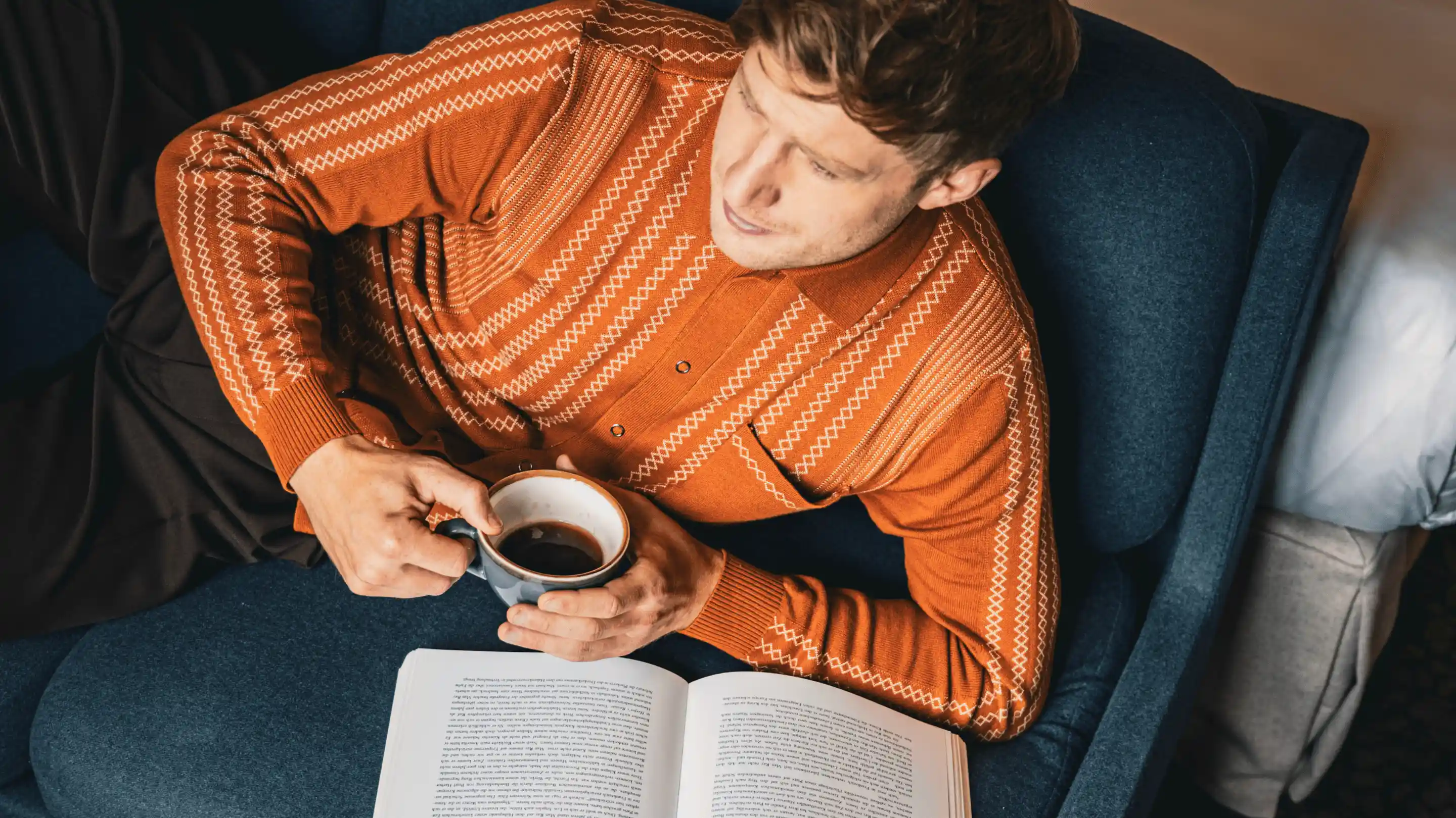 Man in an orange patterned sweater lying on a blue chair, holding a cup of coffee, and reading an open book.