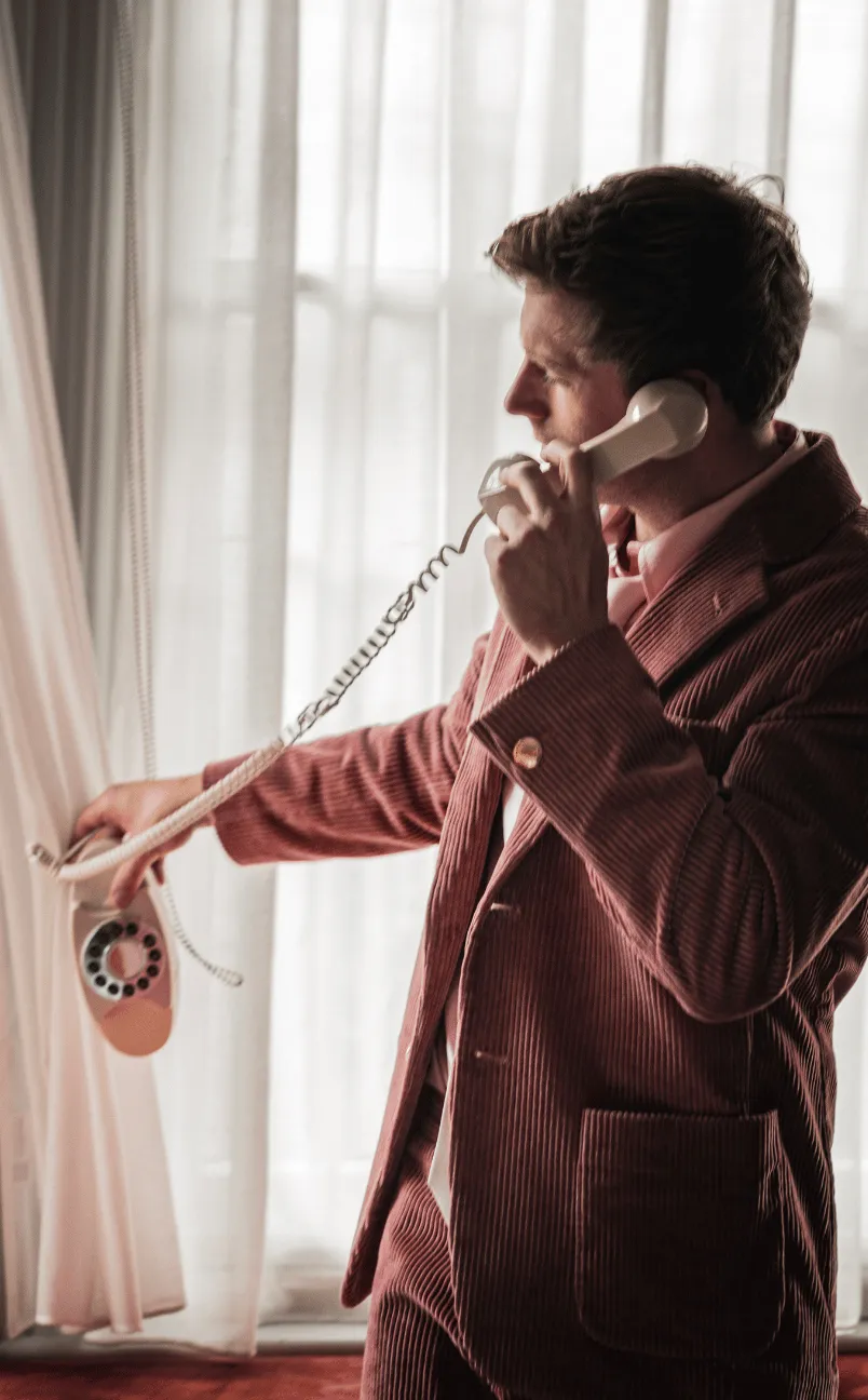 Man in a brown corduroy suit holding a rotary dial phone handset near his ear while reaching for the phone base.