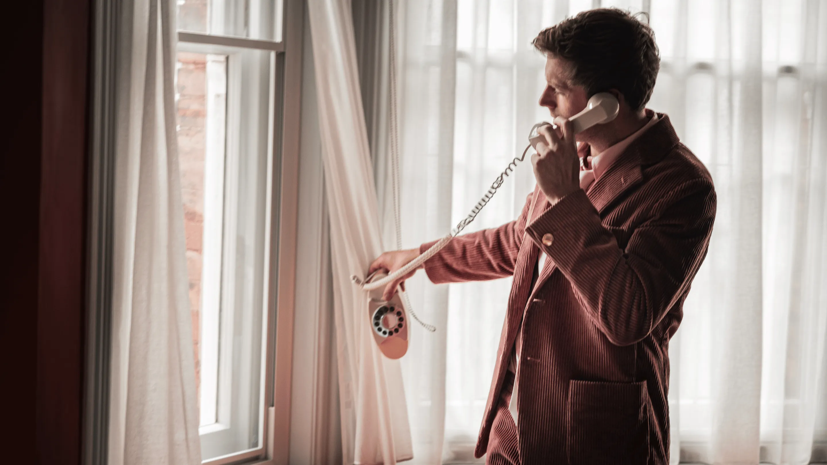 Man in a brown corduroy suit using a beige rotary phone by a window with light curtains.