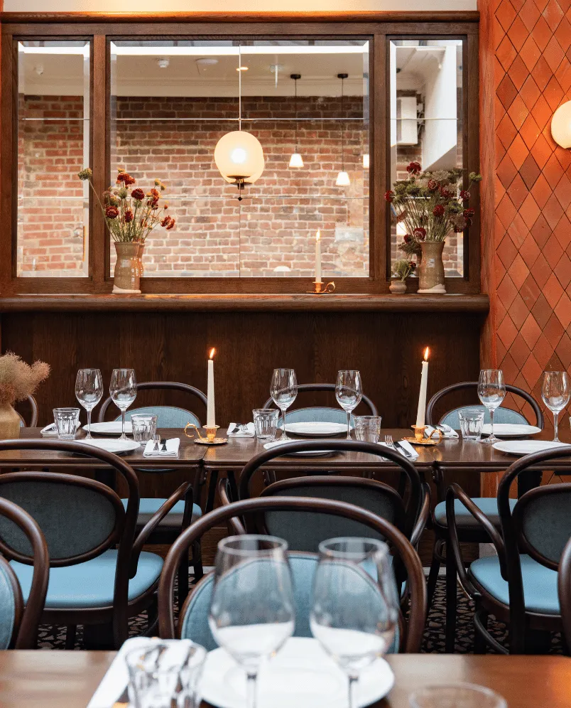 Elegant restaurant table set with wine glasses, plates, lit candles, and blue cushioned chairs in front of a window and brick wall.