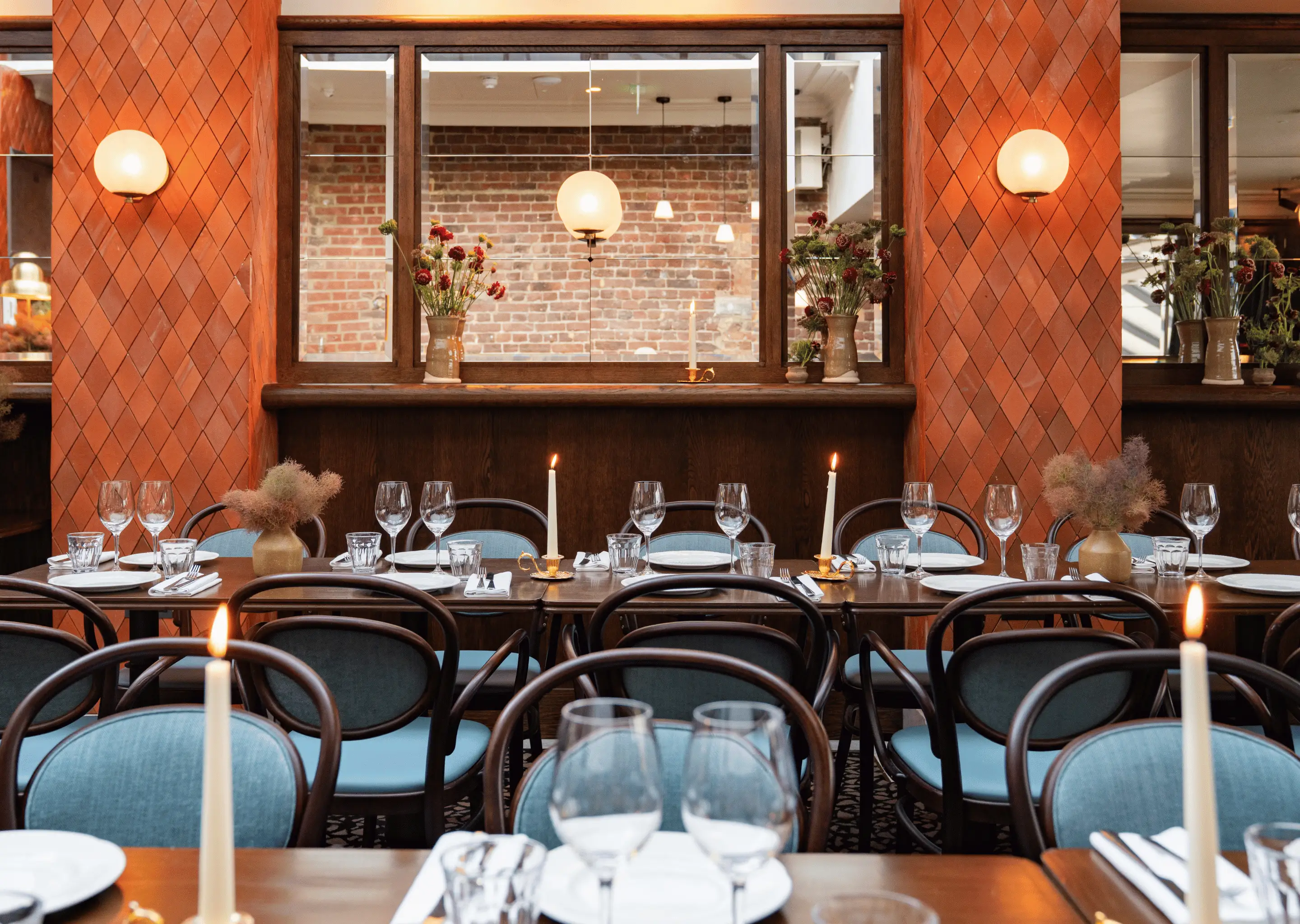 Dining table set with plates, glasses, cutlery, and lit candles in a warm, elegant restaurant interior with blue chairs and brick wall reflections.