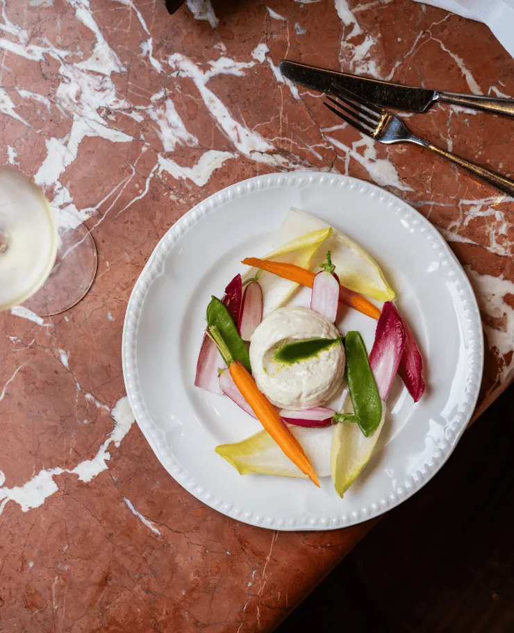 Plate with a round serving of soft cheese surrounded by colorful fresh vegetables including carrot sticks, radicchio leaves, snap peas, and radish slices on a marble table.