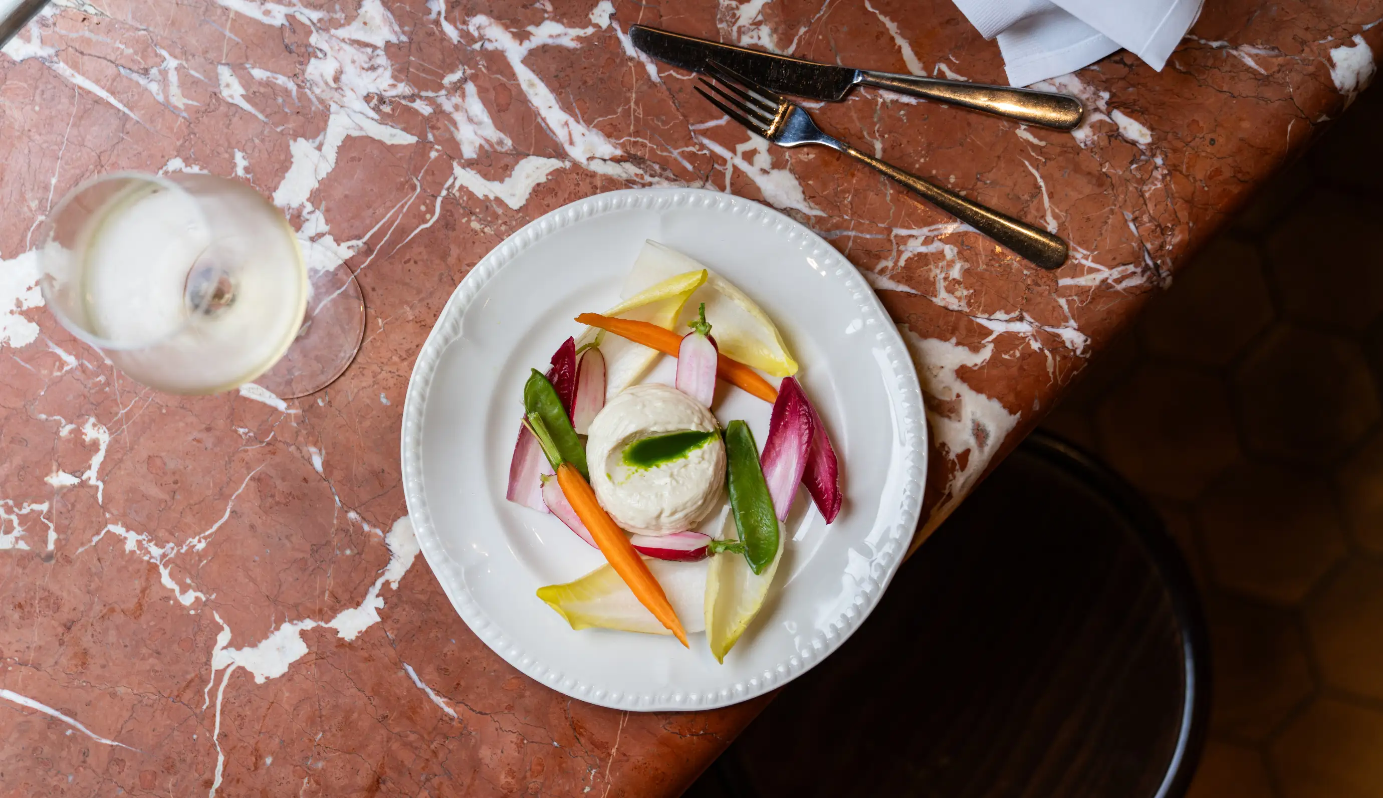 Gourmet plate with a round dollop of creamy cheese topped with green sauce, surrounded by colorful vegetable garnishes including carrots, radish slices, endive leaves, and snap peas on a white plate, next to a glass of white wine on a marble table.