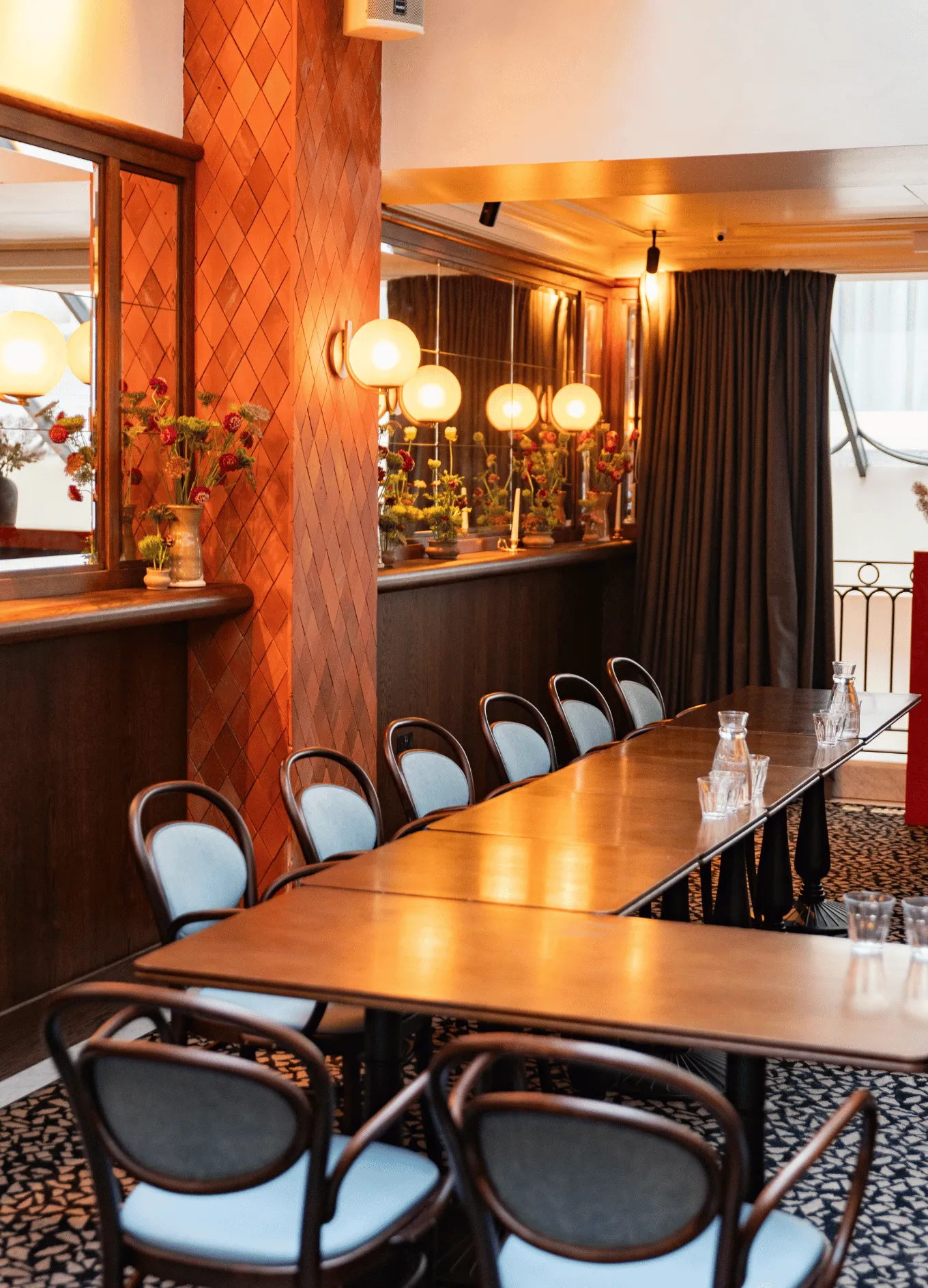Cozy hotel meeting room with long wooden tables, light blue cushioned chairs, warm wall lamps, and floral decorations on a mirrored ledge.